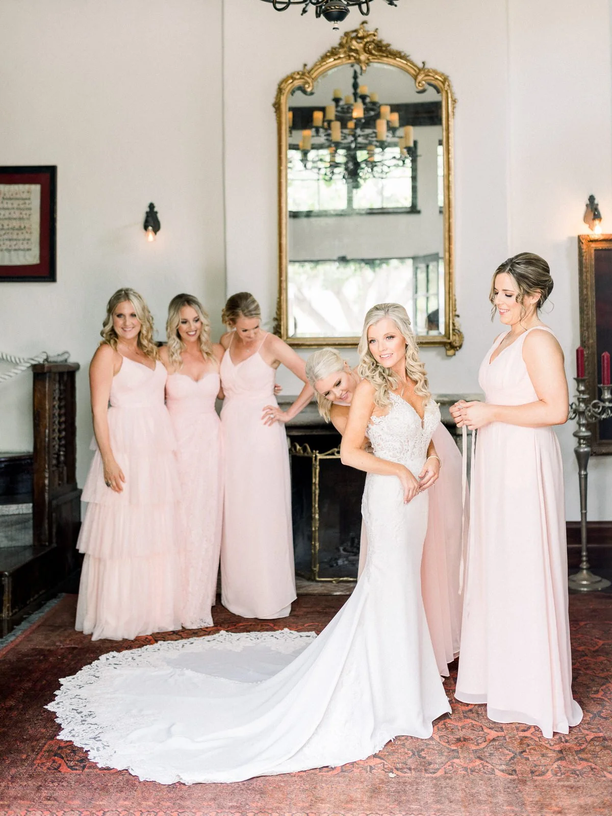 A bride in a white wedding gown with pearl accents is surrounded by five bridesmaids wearing pink dresses. One bridesmaid helps adjust the bride's dress. The setting is an elegantly decorated room with a large ornate mirror, a chandelier, and vintage furniture.