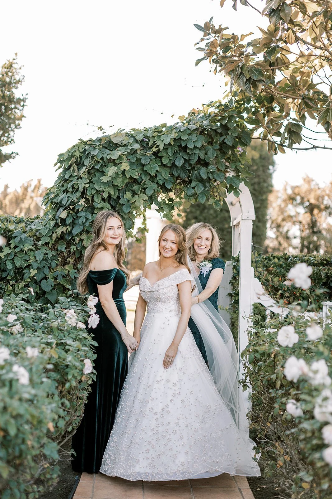Three women, including a bride in a white wedding gown, standing under an arbor covered with green foliage, smiling for a photo in a garden setting.