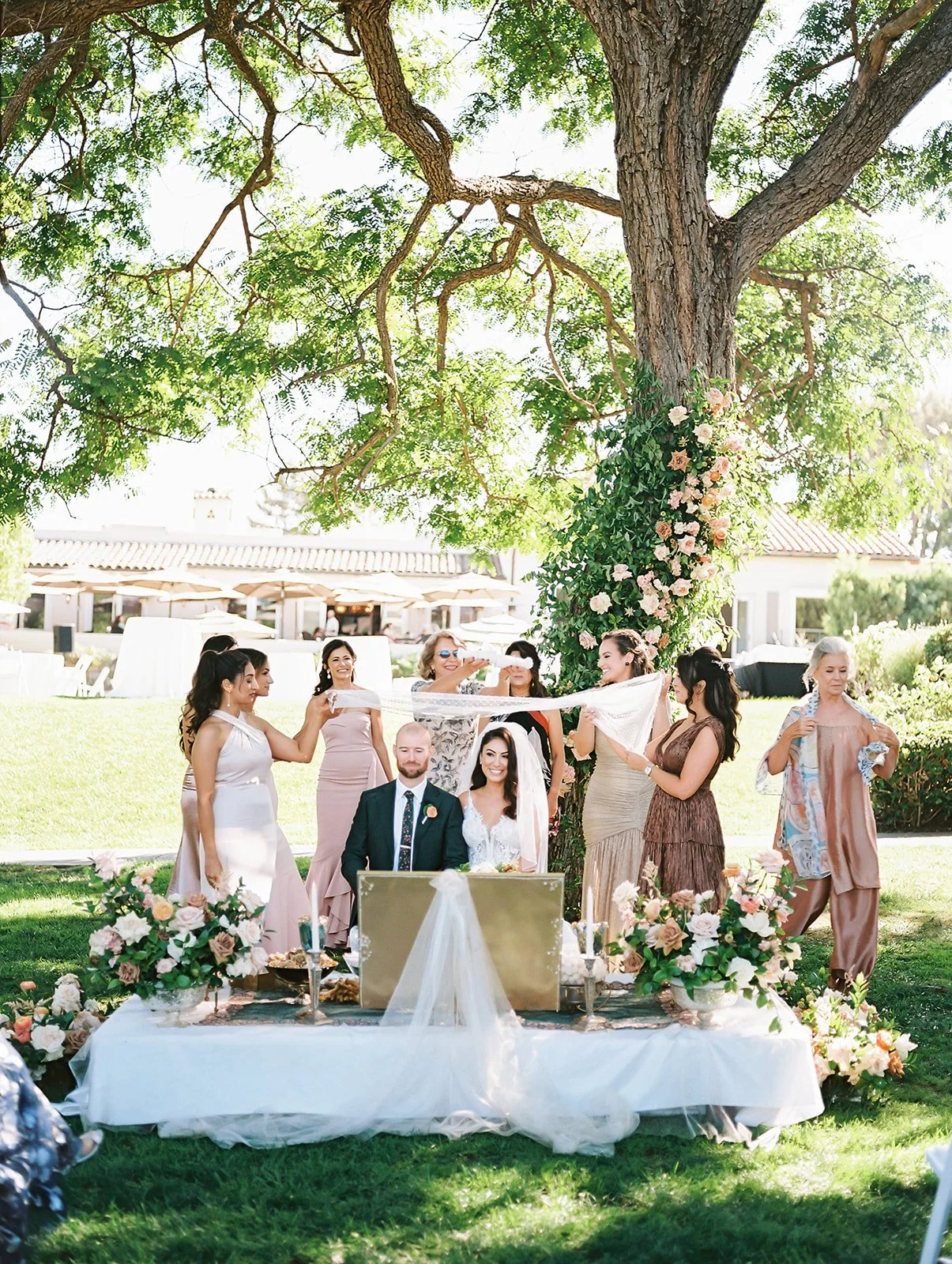 A wedding celebration outdoors under a large tree with a decorated table and bride and groom seated, surrounded by women in pastel dresses, with one woman holding a veil above the couple, in a sunny garden setting.