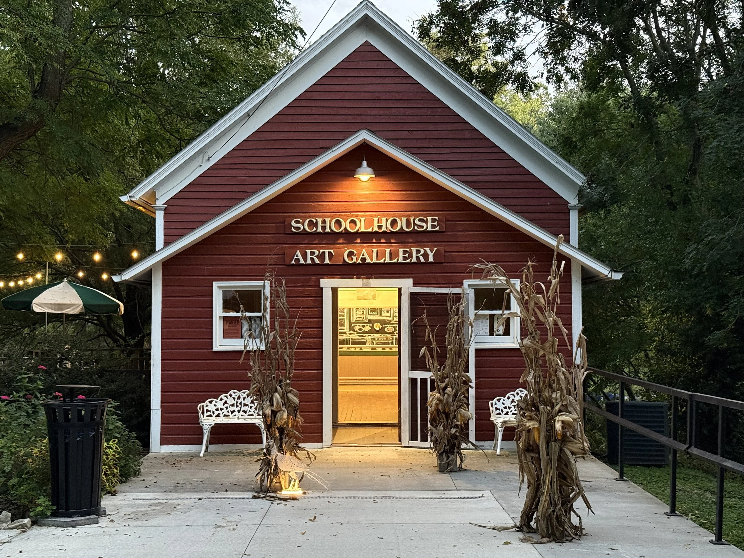 Red building with a sign that reads 'Schoolhouse Art Gallery' illuminated by a light above it, surrounded by trees and autumn decorations, including dried cornstalks and outdoor seating.
