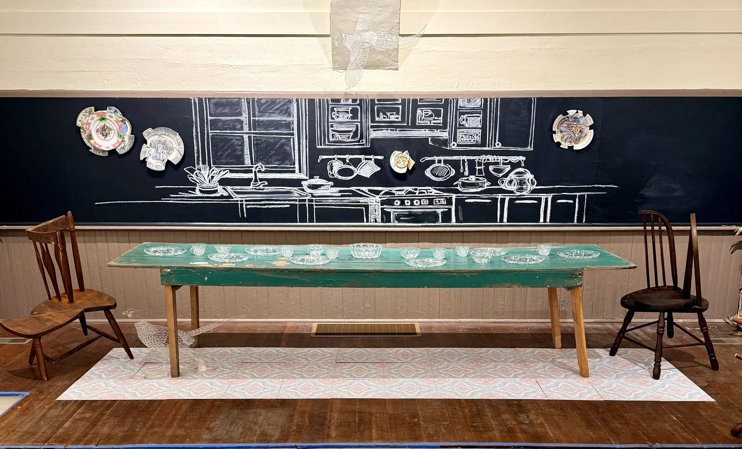 A restaurant interior with a large black chalkboard wall featuring a white chalk drawing of a kitchen scene, decorated with colorful ceramic plates. A wooden table with glass dishes is in front, surrounded by vintage wooden chairs and one dark wooden chair, with a patterned rug underneath.