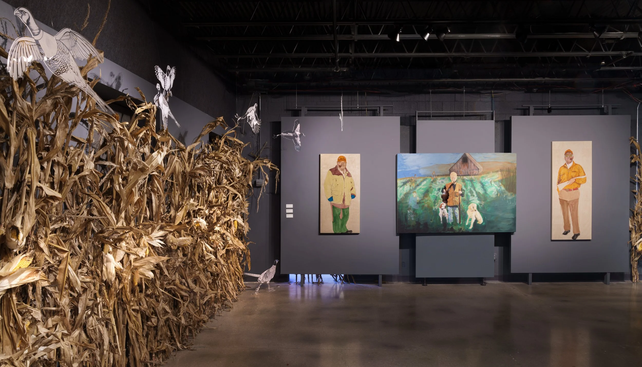 An art exhibit with paintings of people, including a man in a yellow jacket and a woman in an orange jacket, and a landscape scene. To the left, there's dried corn stalks and hanging paper cutouts of birds.