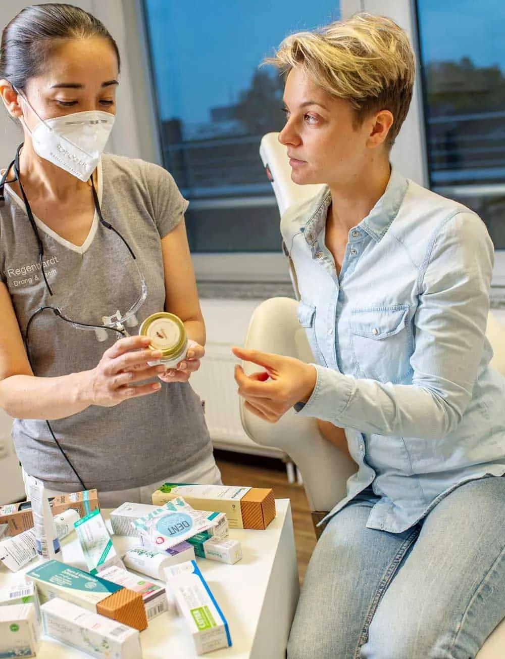 Zwei Frauen unterhalten sich in einem Krankenhauszimmer, eine trägt eine Schutzmaske, während die andere ohne Maske ist, neben einem Tisch mit verschiedenen Medikamenten.