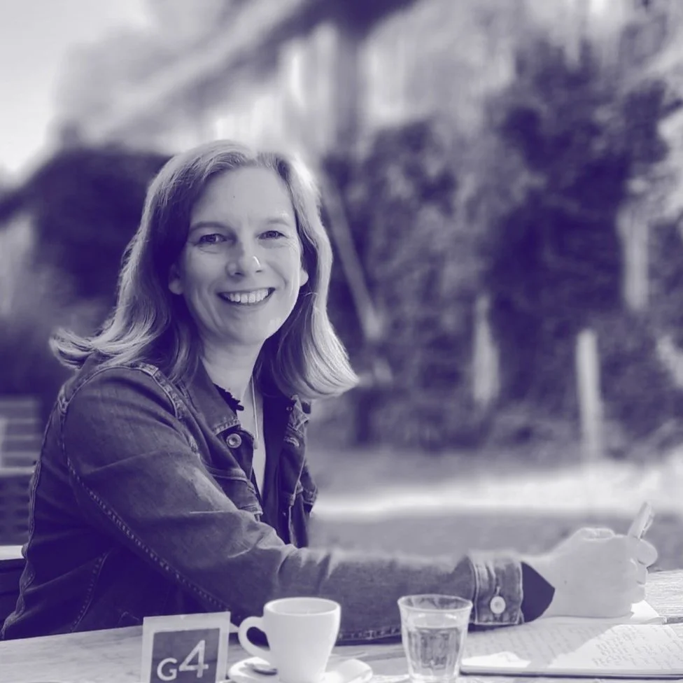 Junge Frau sitzt an einem Tisch im Freien, lächelt in die Kamera, schreibt etwas und hat eine Tasse, ein Glas Wasser und eine Notiz vor sich.