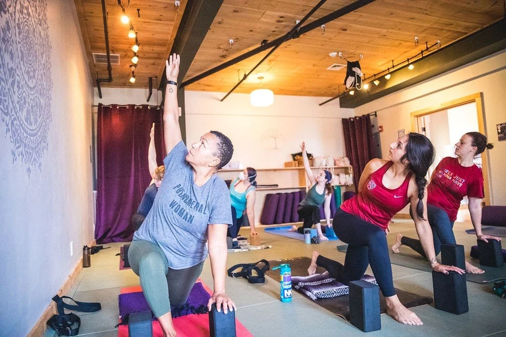 Students practicing a twisting pose using blocks in a yoga studio with natural lighting.