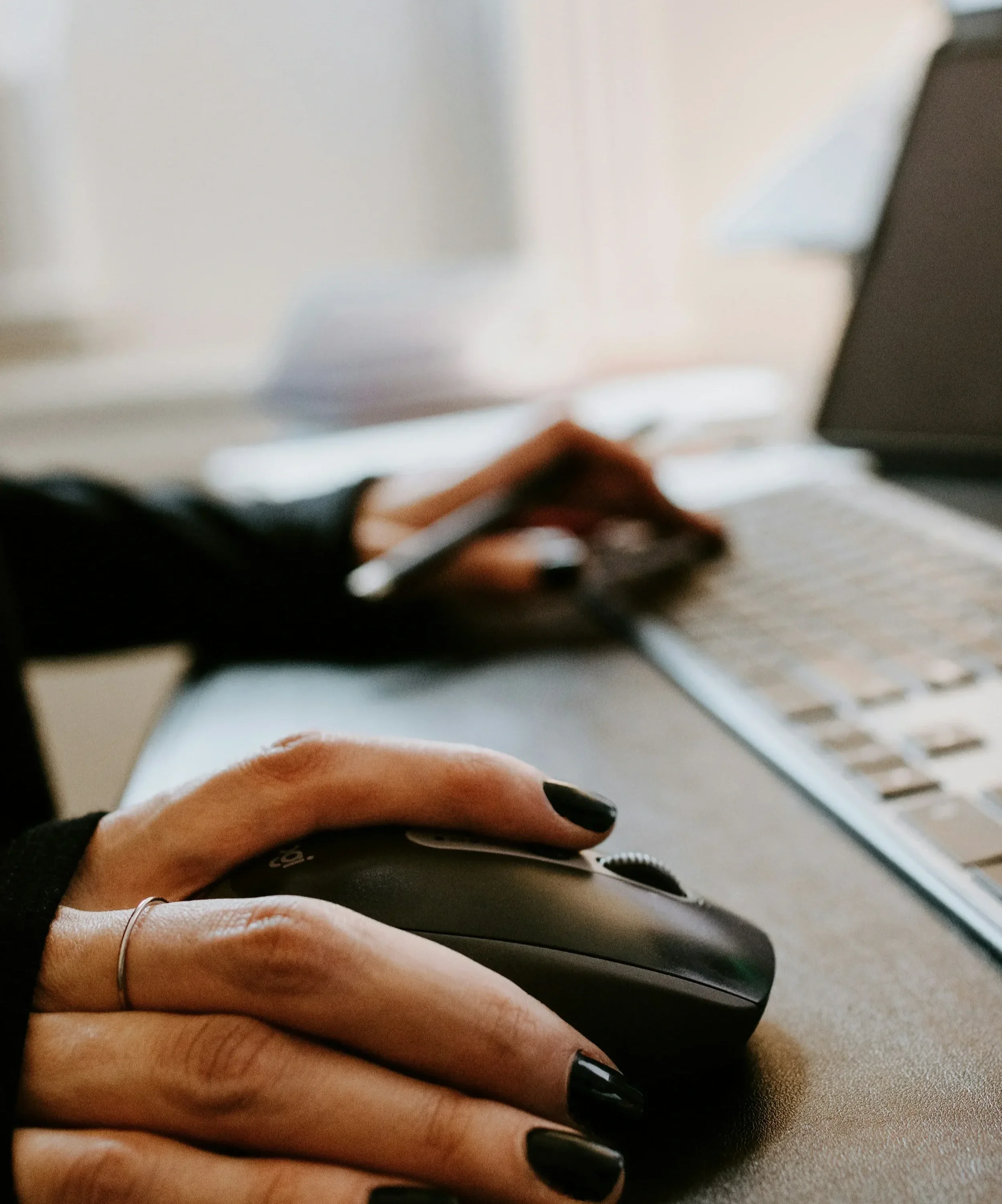 Close-up of a person's hand with black painted nails using a computer mouse on a brown surface, with a blurred keyboard nearby.