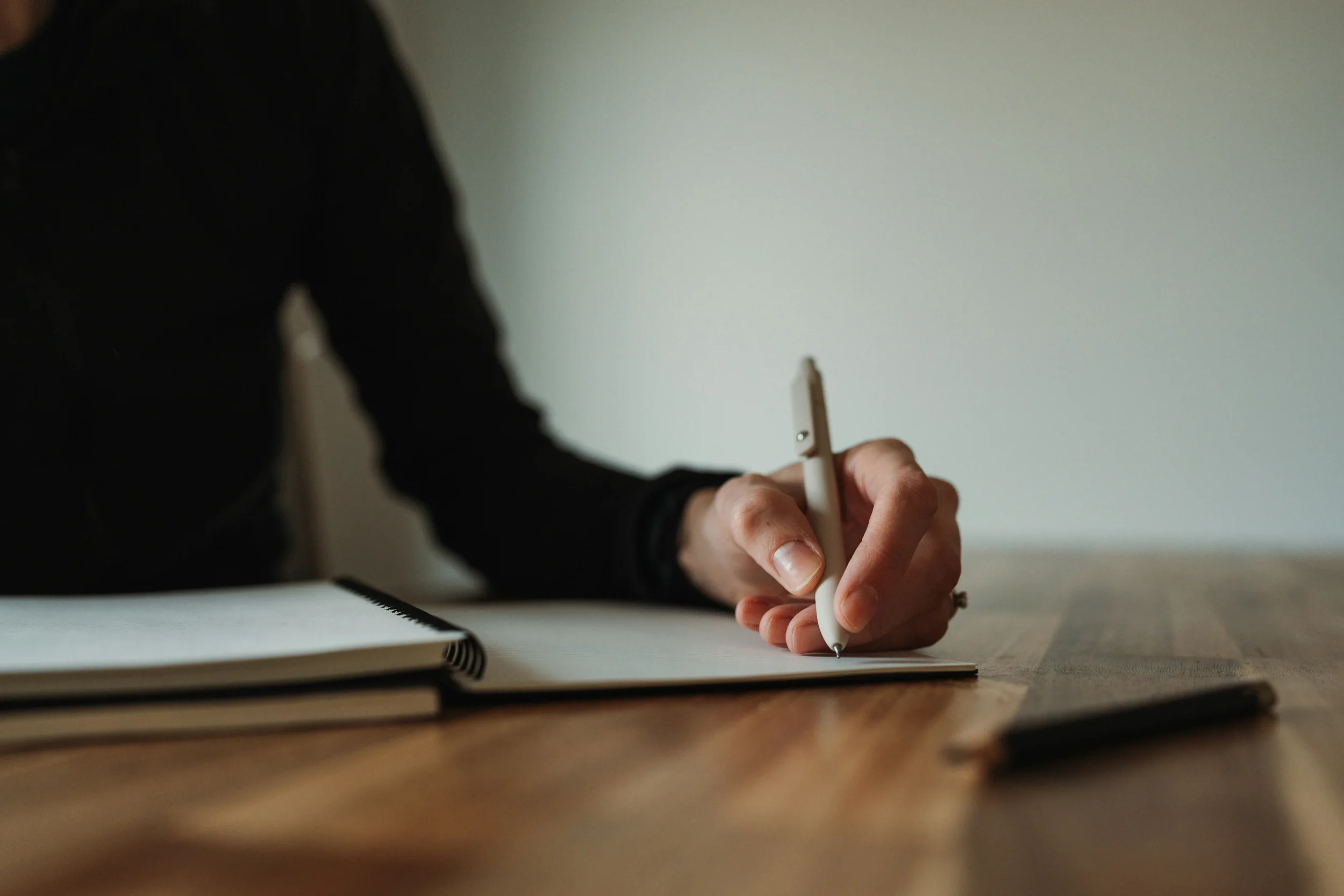 Person writing with a white pen on paper, with notebooks or folders on a wooden table.