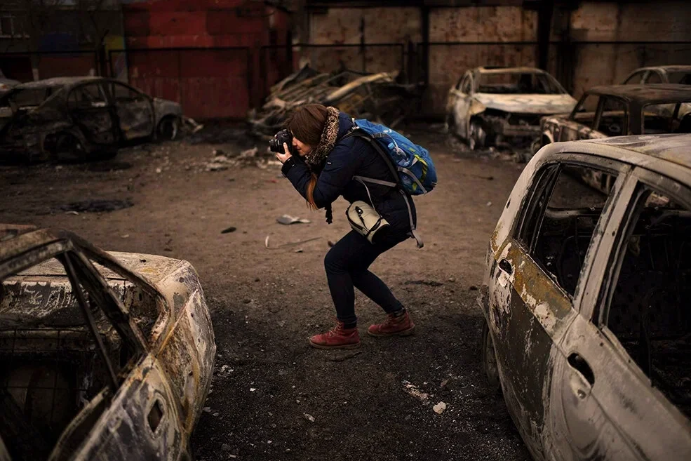 A woman with a backpack crouches and takes a photograph among numerous burned and damaged cars in a destructive scene.