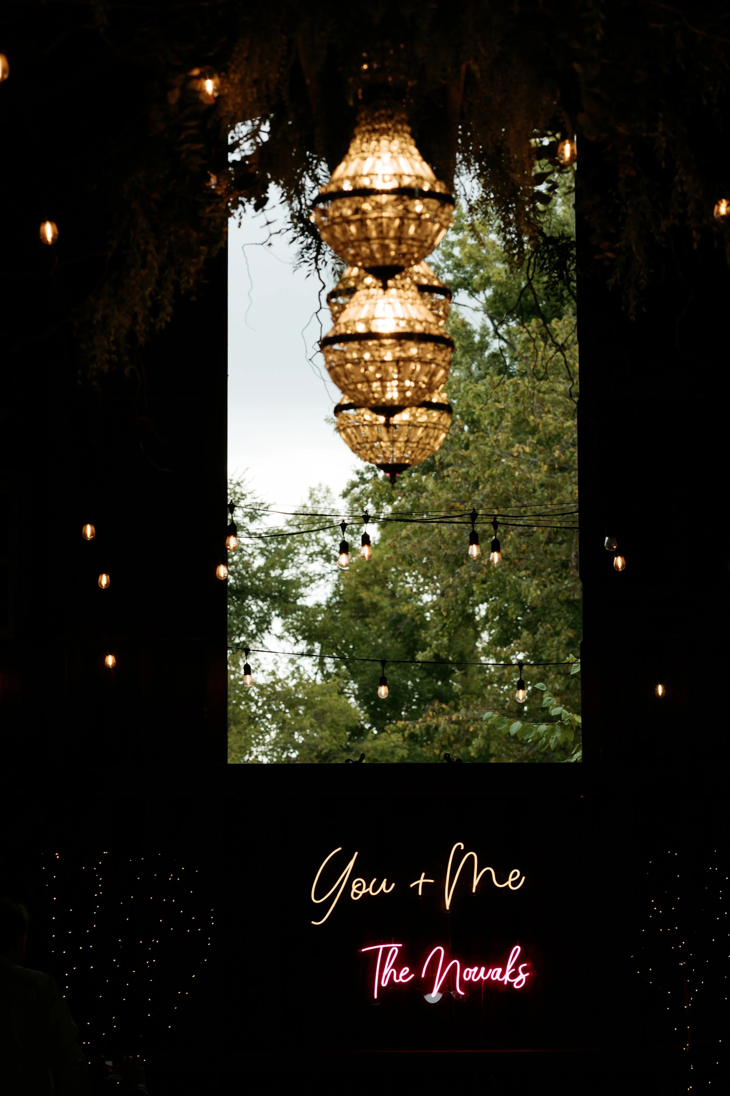 Neon sign reading 'You + Me' and 'The Nowaks' against a dark background, with hanging string lights and a window showing trees outside.