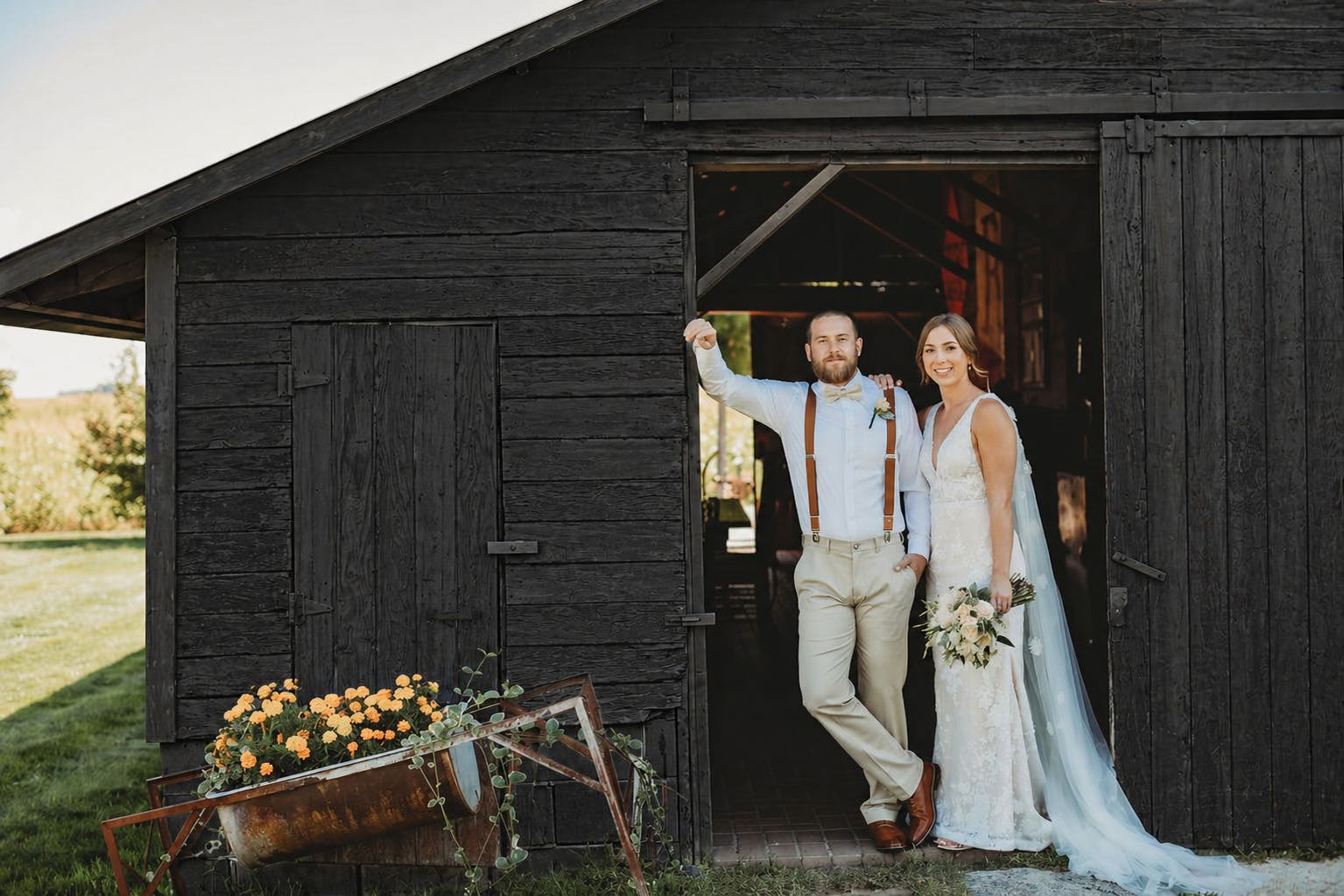 A couple dressed in wedding attire standing in a rustic wooden barn door opening outdoors during daytime, with a smiling bride holding a bouquet of flowers and a groom with suspenders leaning against the barn frame.