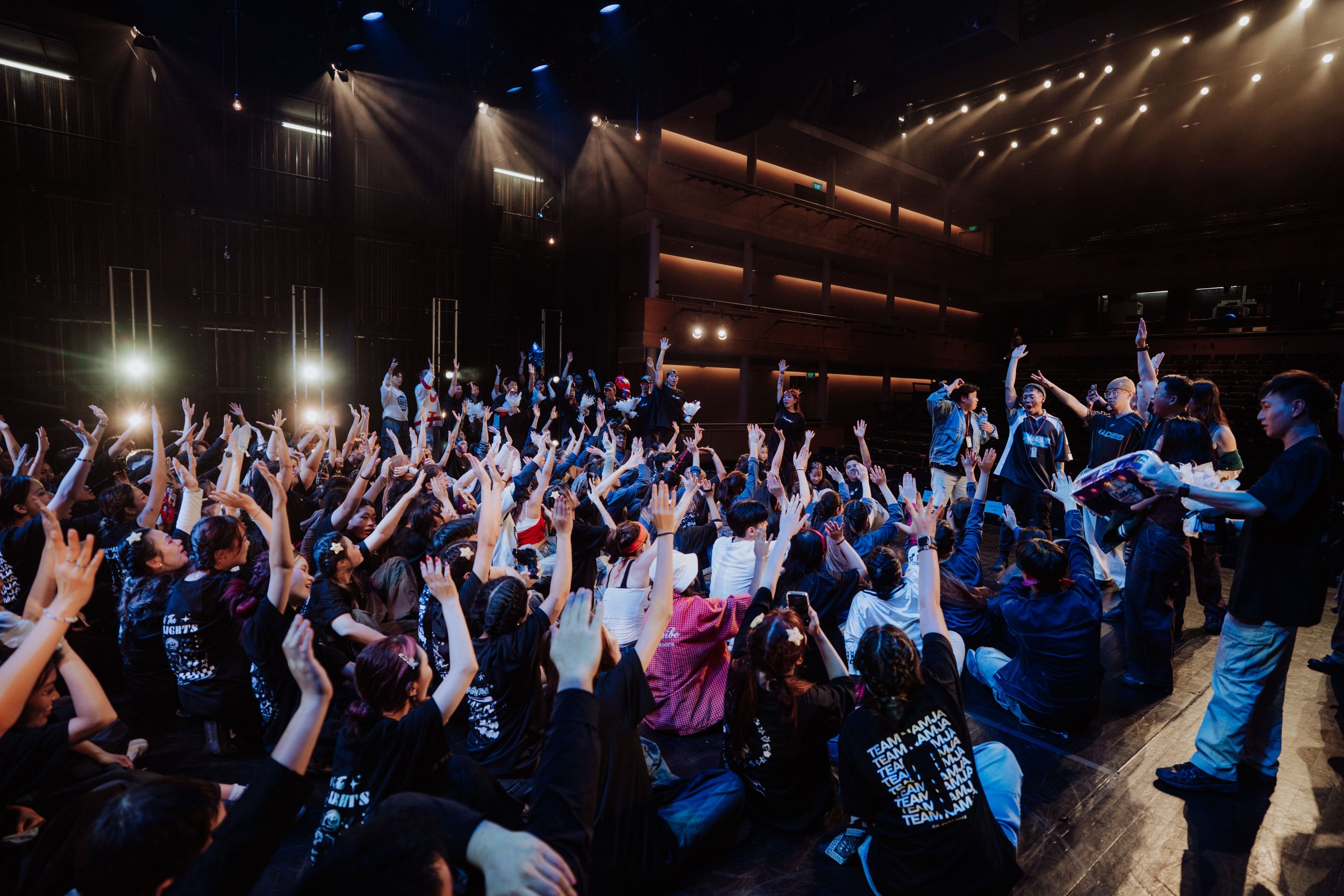 A large crowd of people at a concert or event with many raising their hands. The stage has bright lights and the venue is an indoor auditorium with multiple levels and balconies.