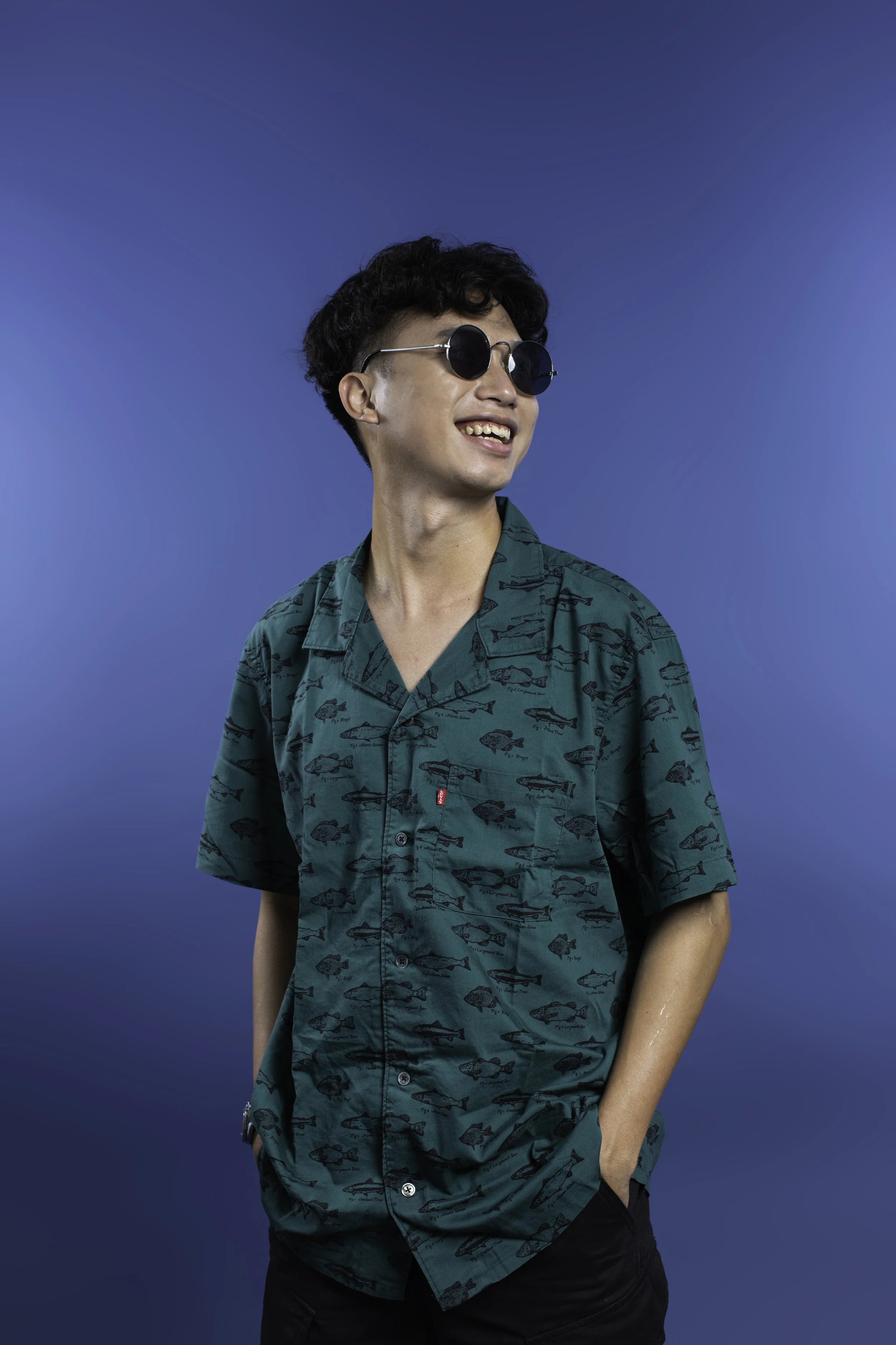 A young man with curly black hair wearing round black sunglasses and a teal short-sleeved button-up shirt with a fish pattern, smiling and leaning slightly to his right against a blue background.