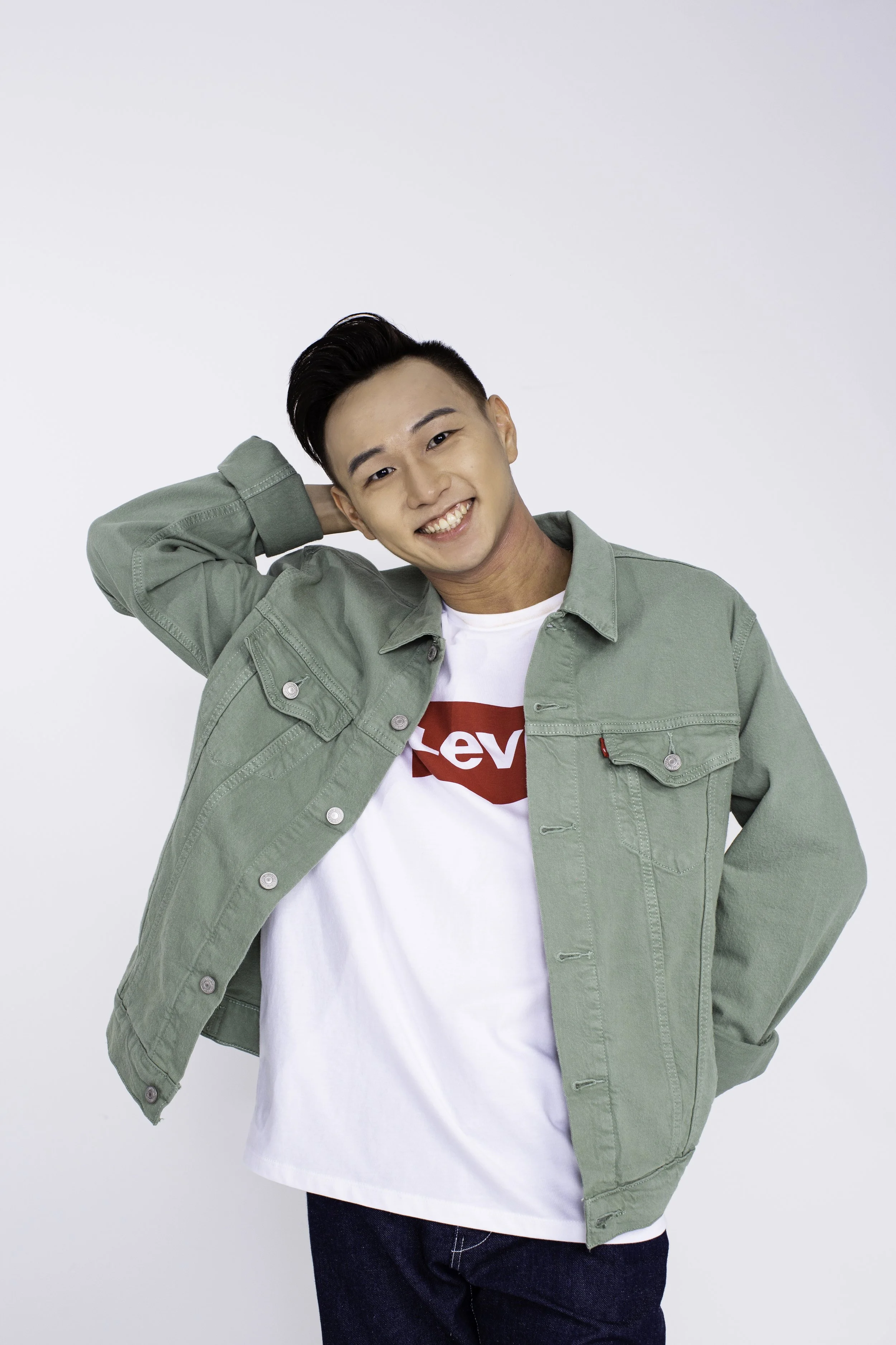 A young man smiling, wearing a green jacket over a white T-shirt with a red Levi's logo, black jeans, and a white shirt underneath, posing with one hand behind his head against a plain white background.