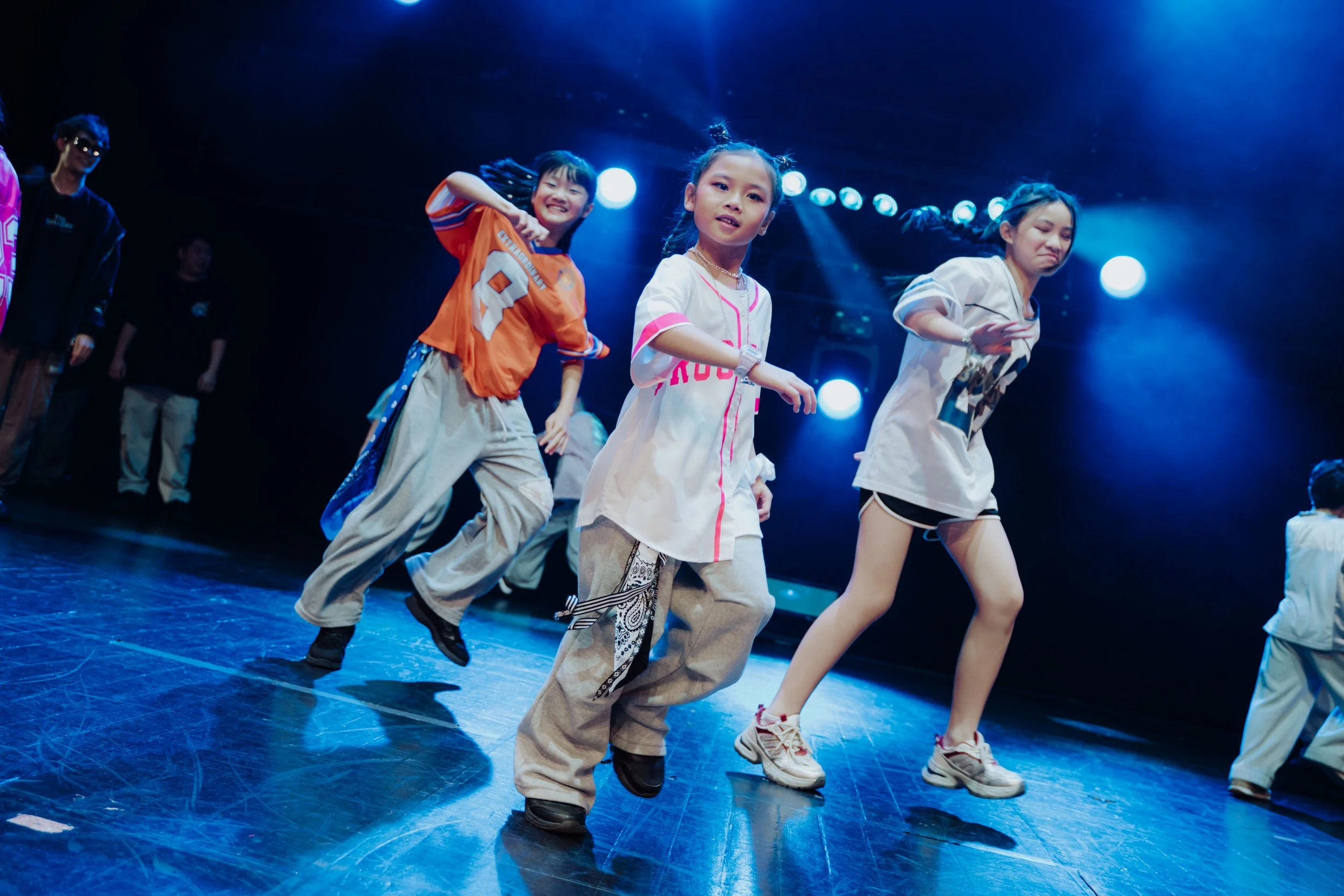 Young girls dancing on stage with colorful outfits and energetic expressions under bright stage lights.
