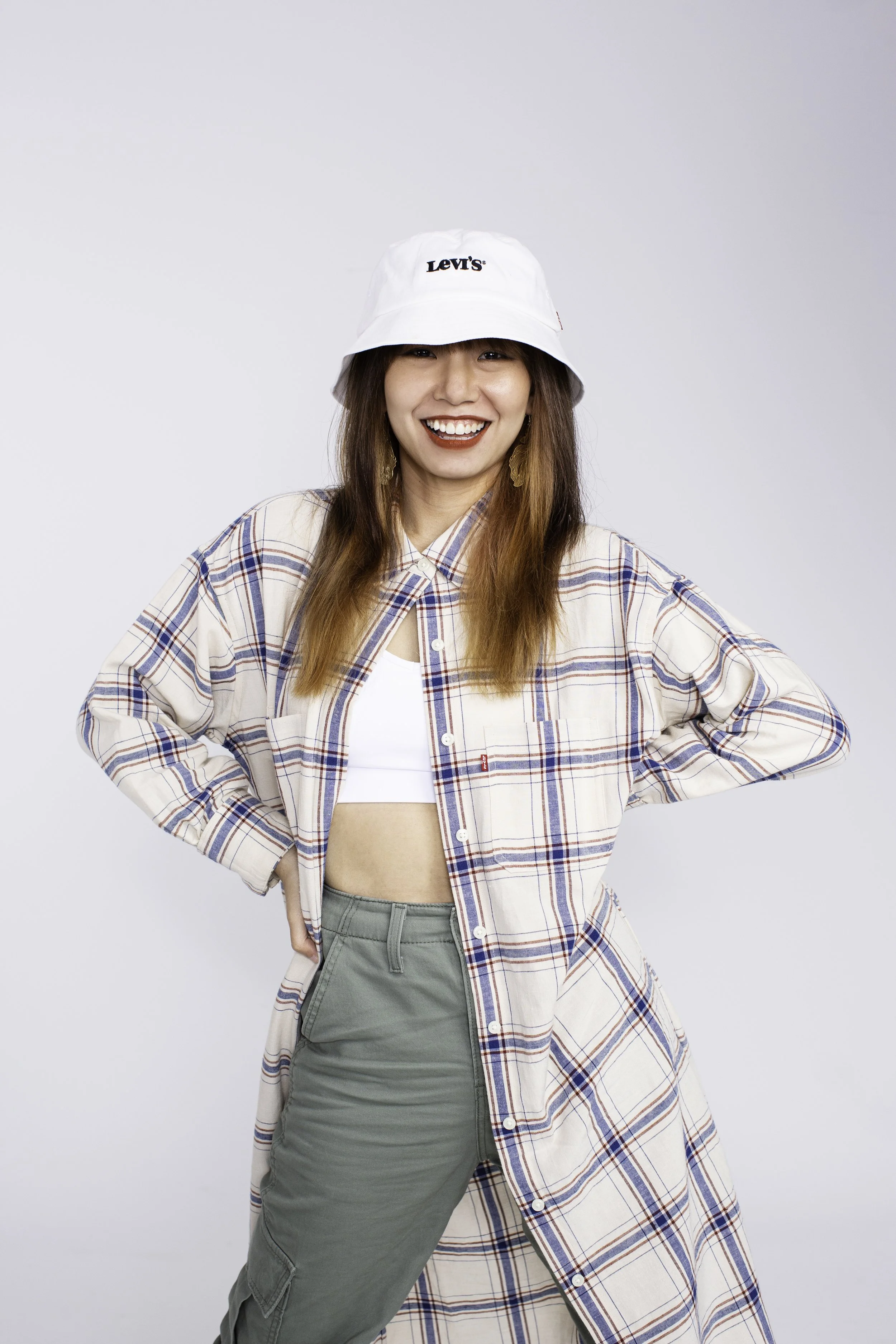 A woman is smiling and wearing a white bucket hat with the Levi's logo, a plaid button-up shirt, and green cargo pants, against a plain white background.