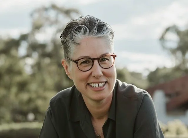 Portrait of a smiling woman with short gray hair, wearing glasses and a dark shirt, outdoors with trees and cloudy sky in the background.