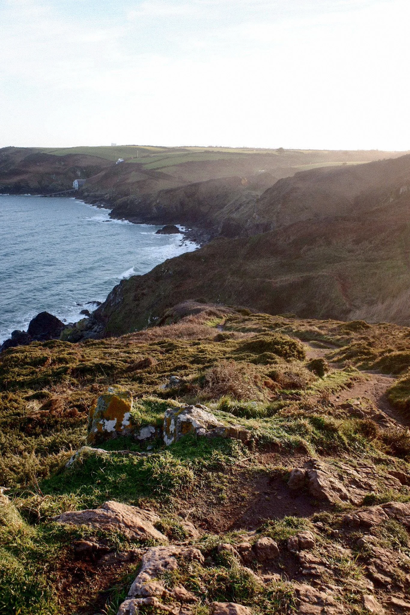 Coastal landscape with rocky cliffs, green grass, and ocean waves at sunset.
