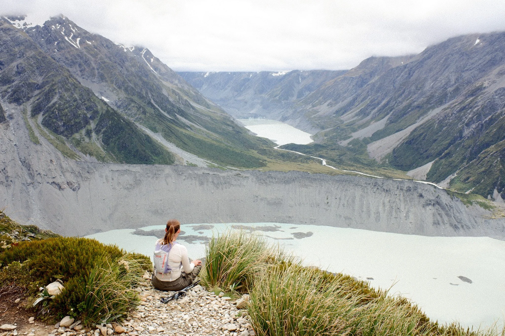 A person sitting on a rocky ledge overlooking a glacial valley with a winding river, surrounded by steep, snow-capped mountains.
