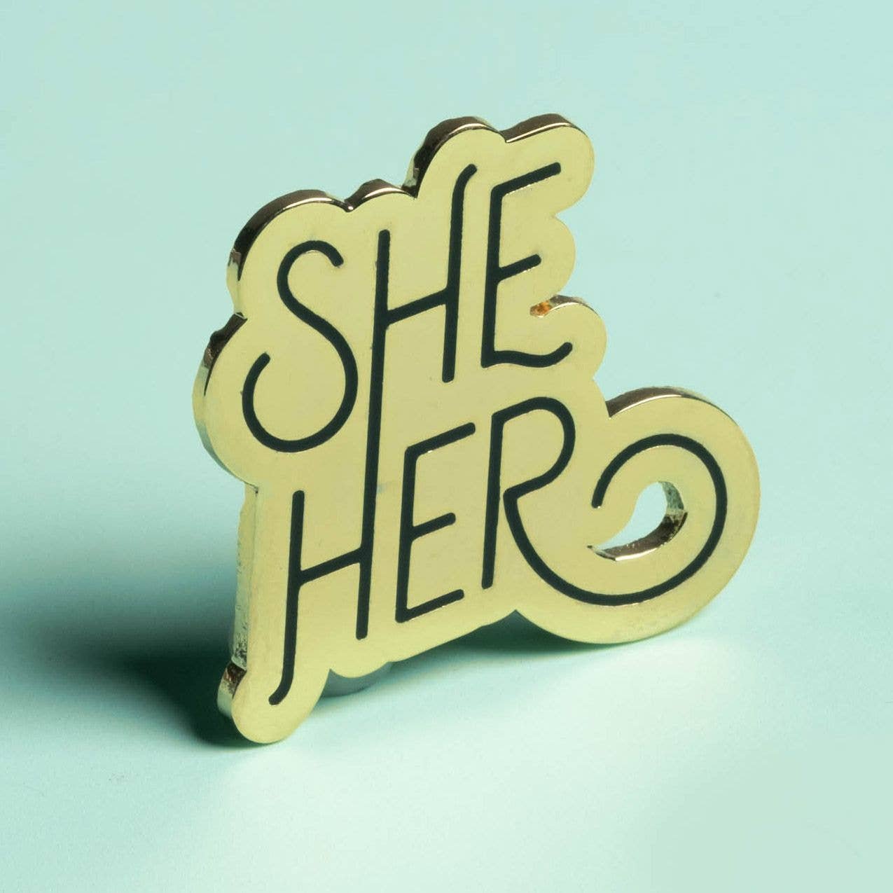 Image Of Gold “She/Her” Pronoun Pin Featuring Gold Metal With Engraved Black Hand-Drawn Type.