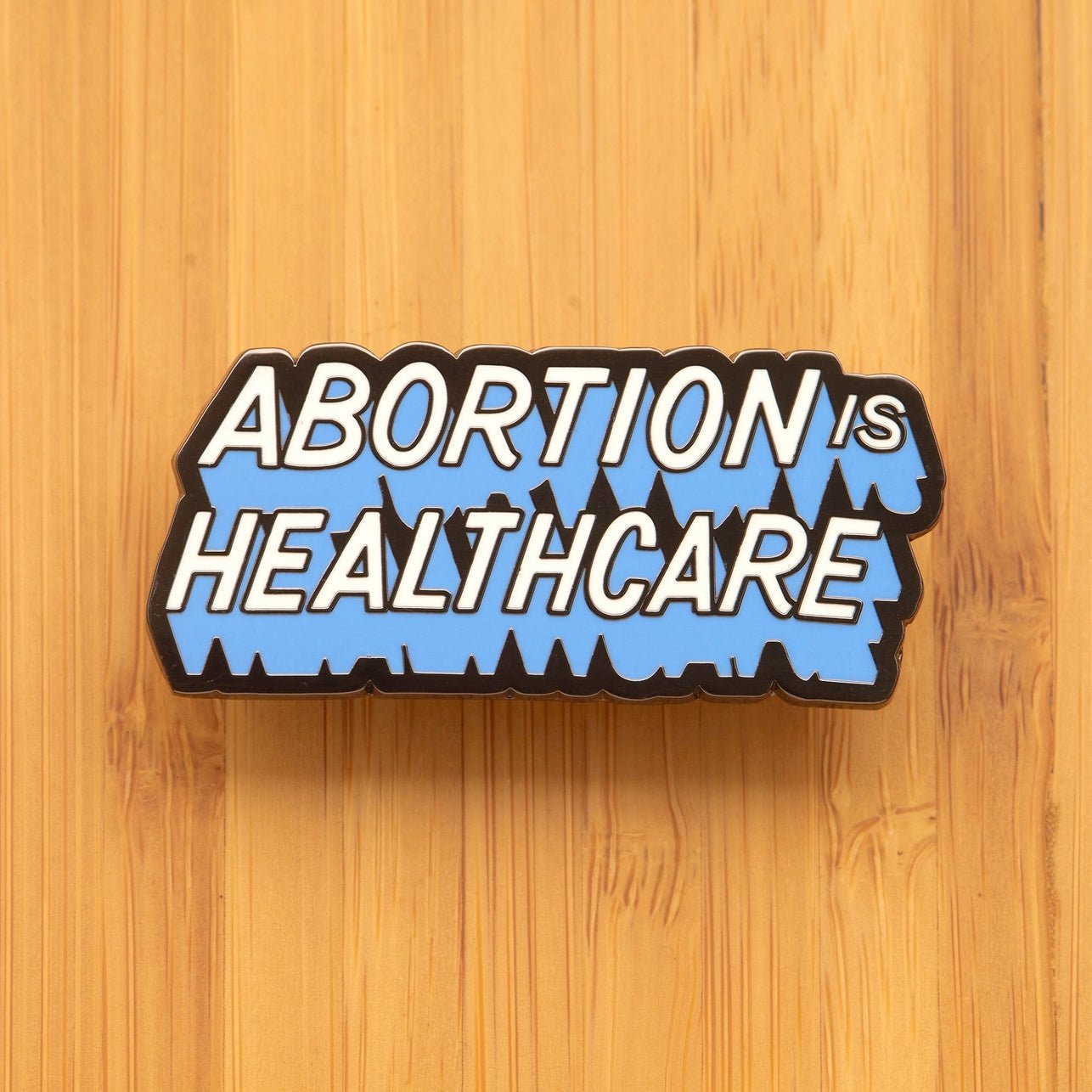 Image Of “Abortion Is Healthcare” Enamel Pin With White, Blue And Black All-Caps Block Type.