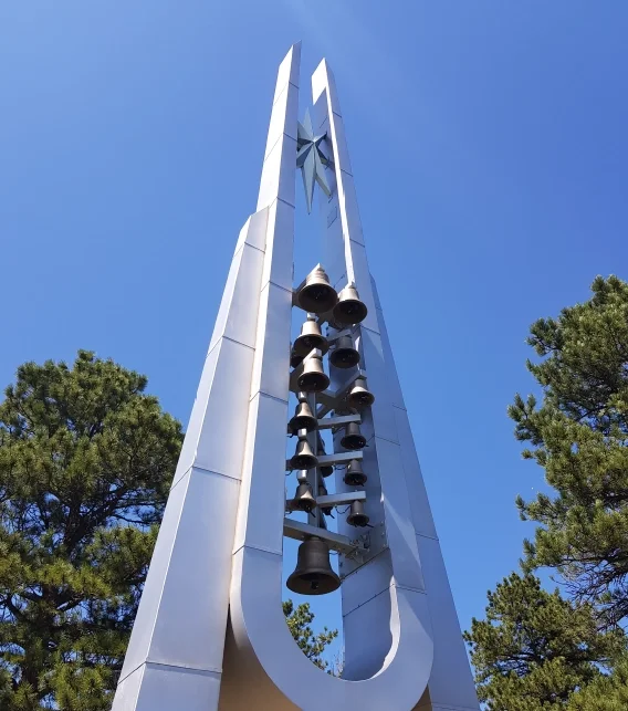 Multiple bronze bells mounted in a modern outdoor carillon tower against a blue sky