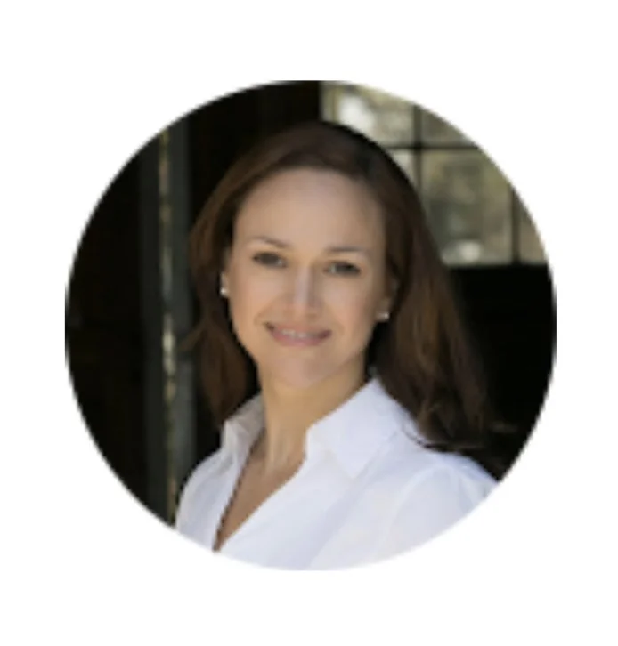 Portrait of a woman with brown hair, wearing a white collared shirt, smiling in front of a window.