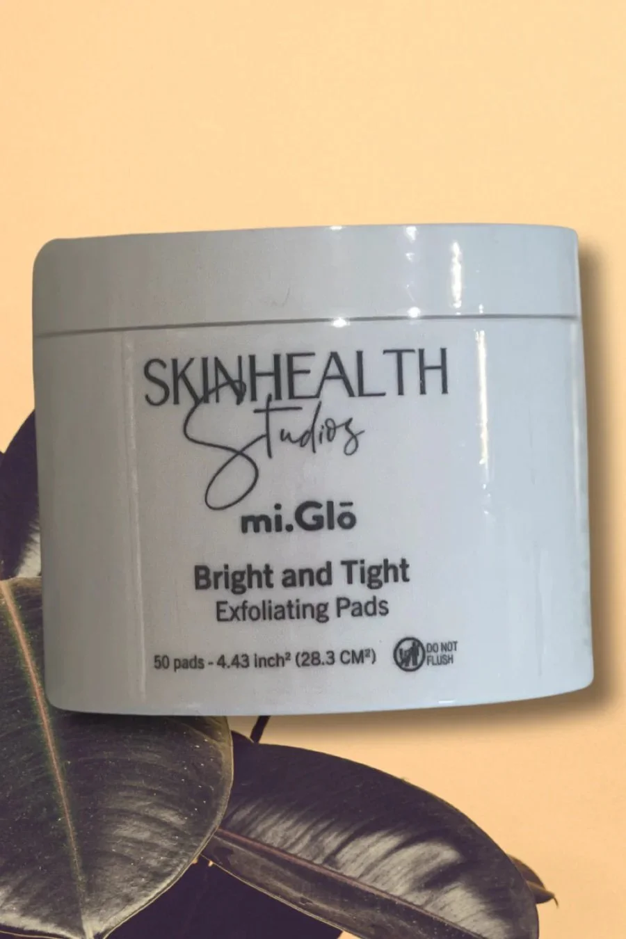 A white container labeled 'SkinHealth Studios mi.Glo Bright and Tight Exfoliating Pads' with a black leafy plant partially visible at the bottom left.