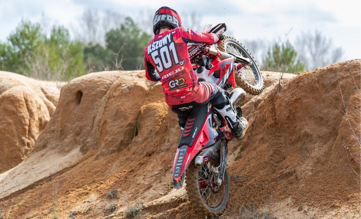 Motocross rider climbing a dirt hill on a red motorcycle in an outdoor setting.