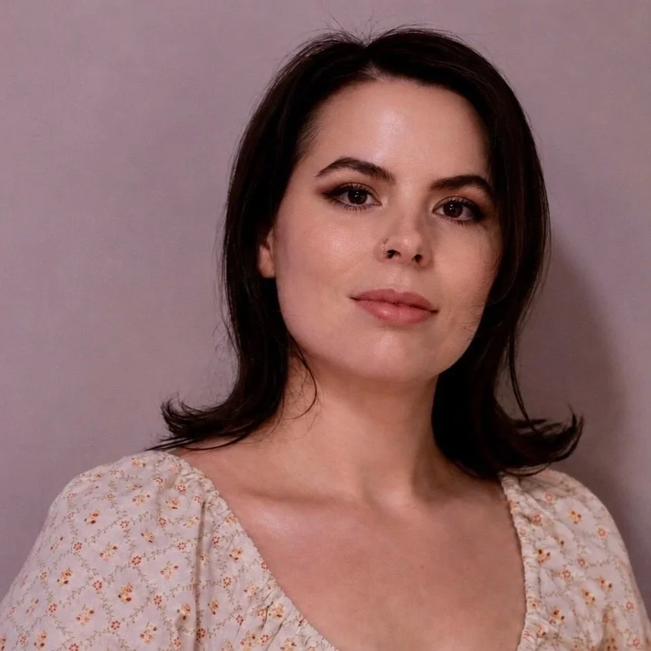A woman with dark hair sitting on a cream-colored sofa, holding a black mug, wearing a black blouse and maroon pants, with a window showing green trees in the background.
