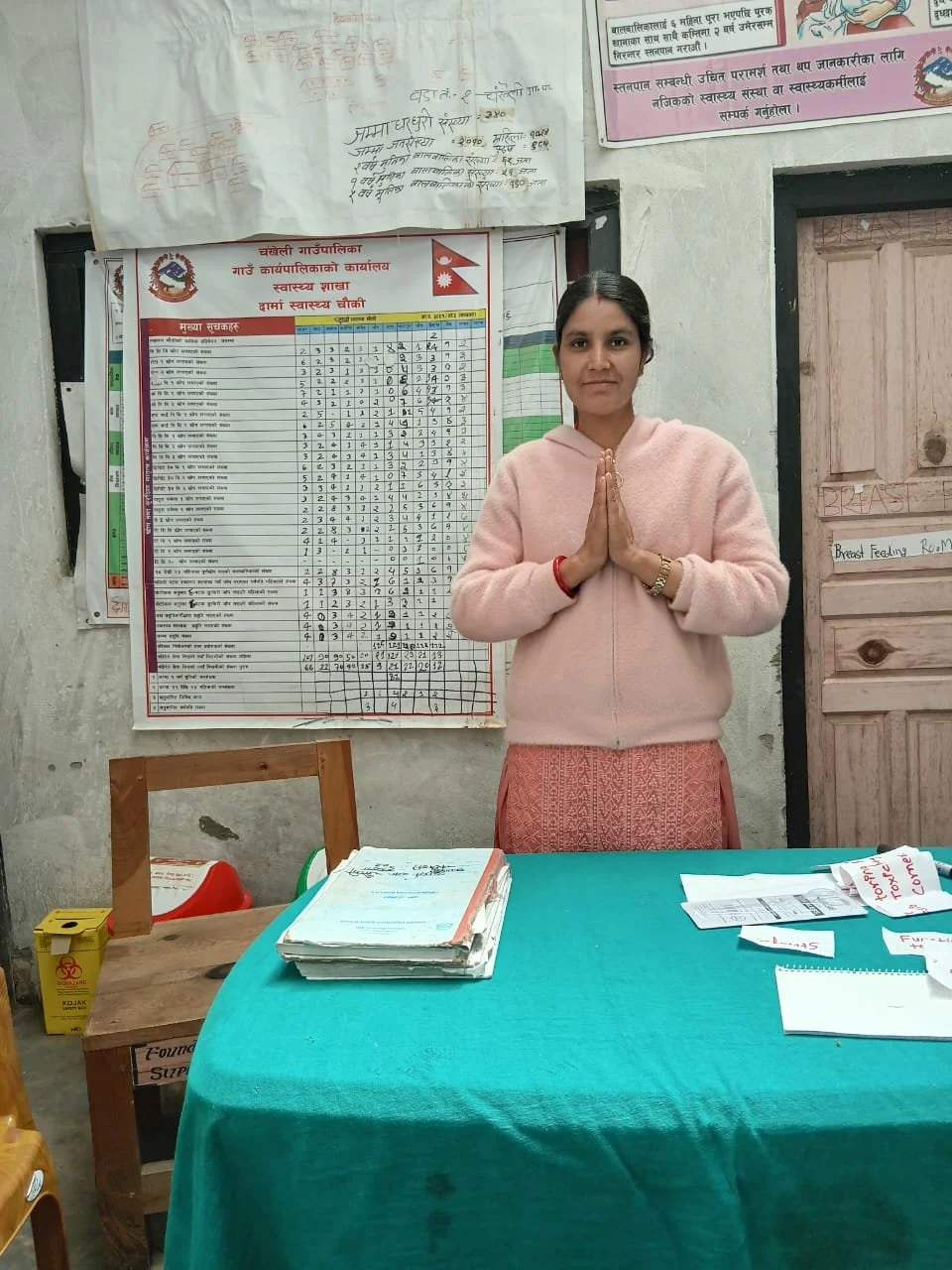 Bandana Jaisi, Auxiliary Nurse Midwife at Darma Health Post in Chankheli Rural Municipality, Humla District, stands at her workstation with hands pressed together in a traditional Nepali greeting. Health data charts and maternal and newborn health ed