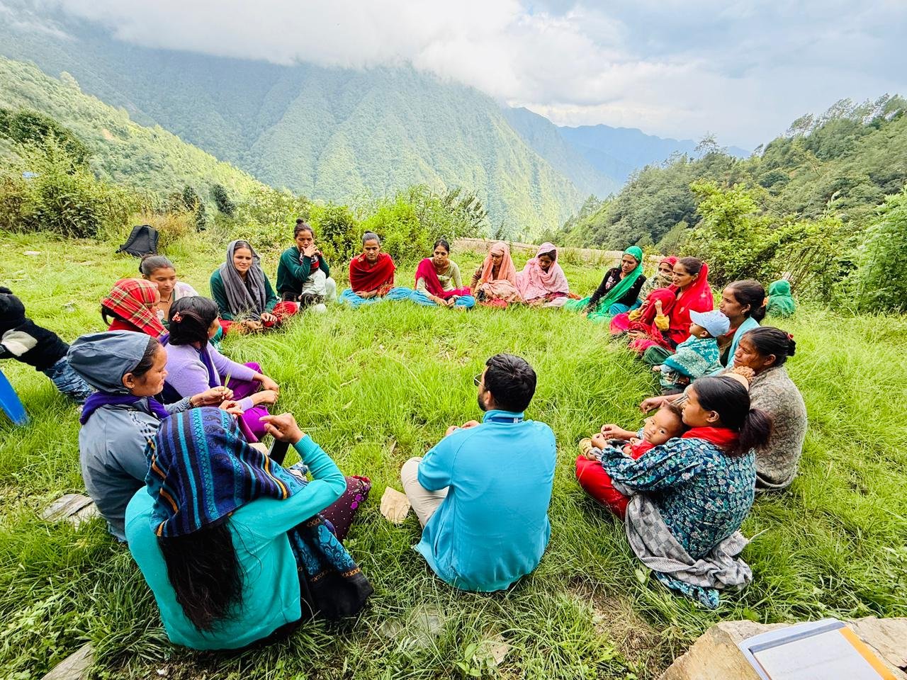 A community outreach worker sits in a circle with a group of mothers and their children on a grassy hillside in Nepal, with green mountains behind them.