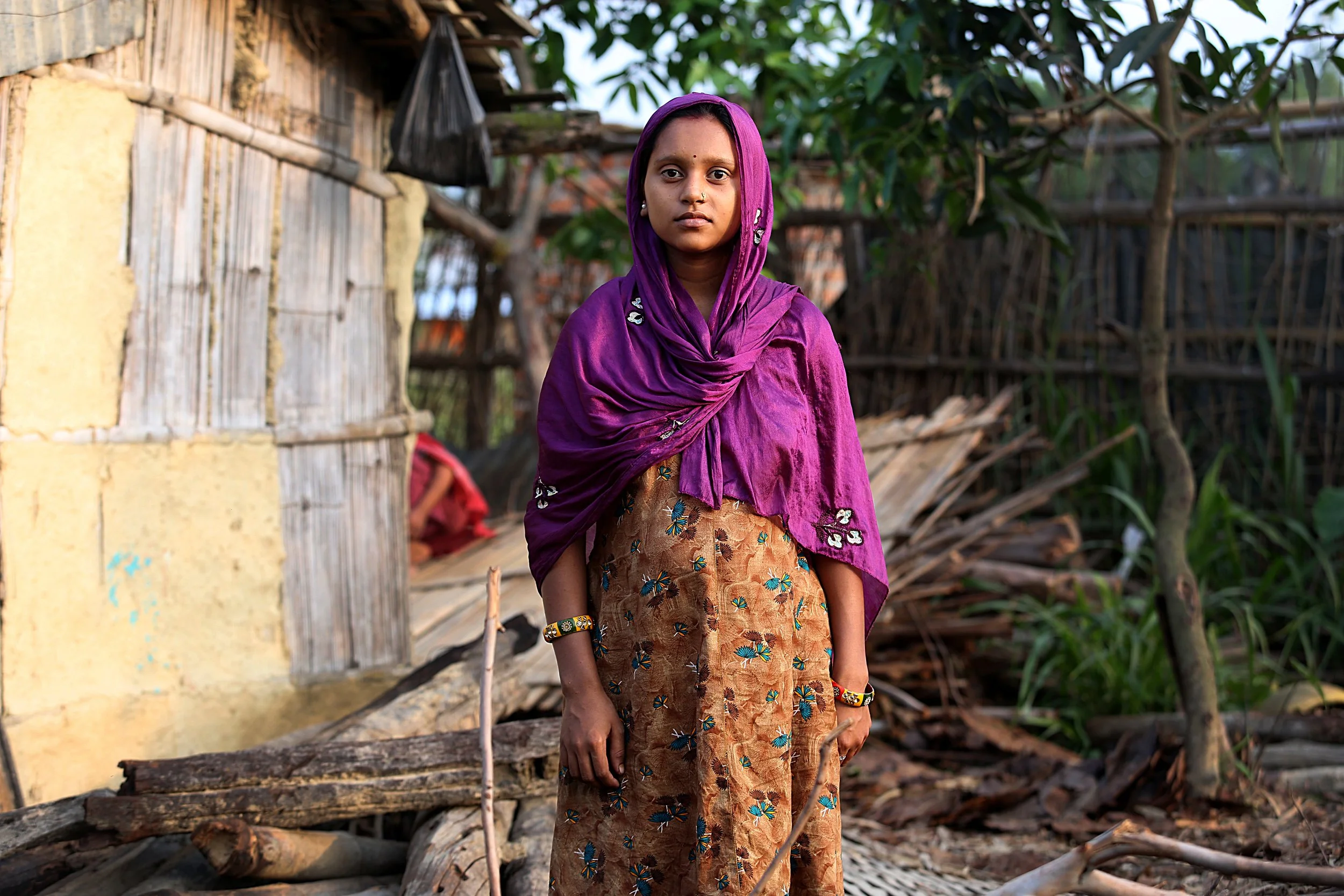 A young woman in a purple headscarf stands and looks toward the camera, outside a home in a rural village.