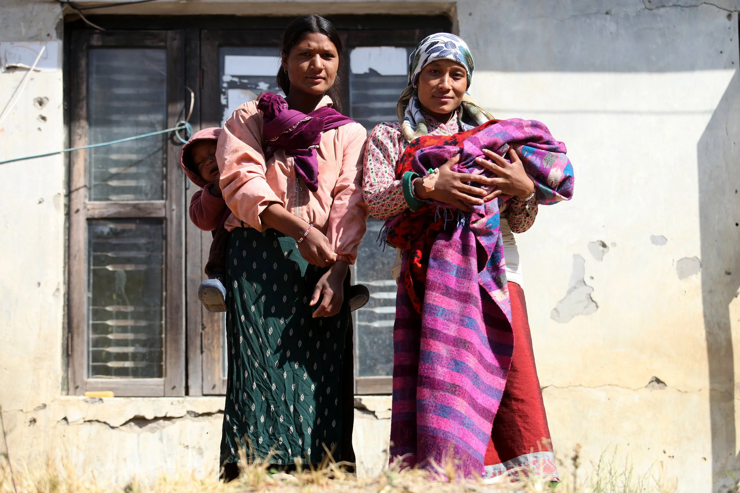 Two mothers in Nepal stand together outside a home, each holding her newborn.