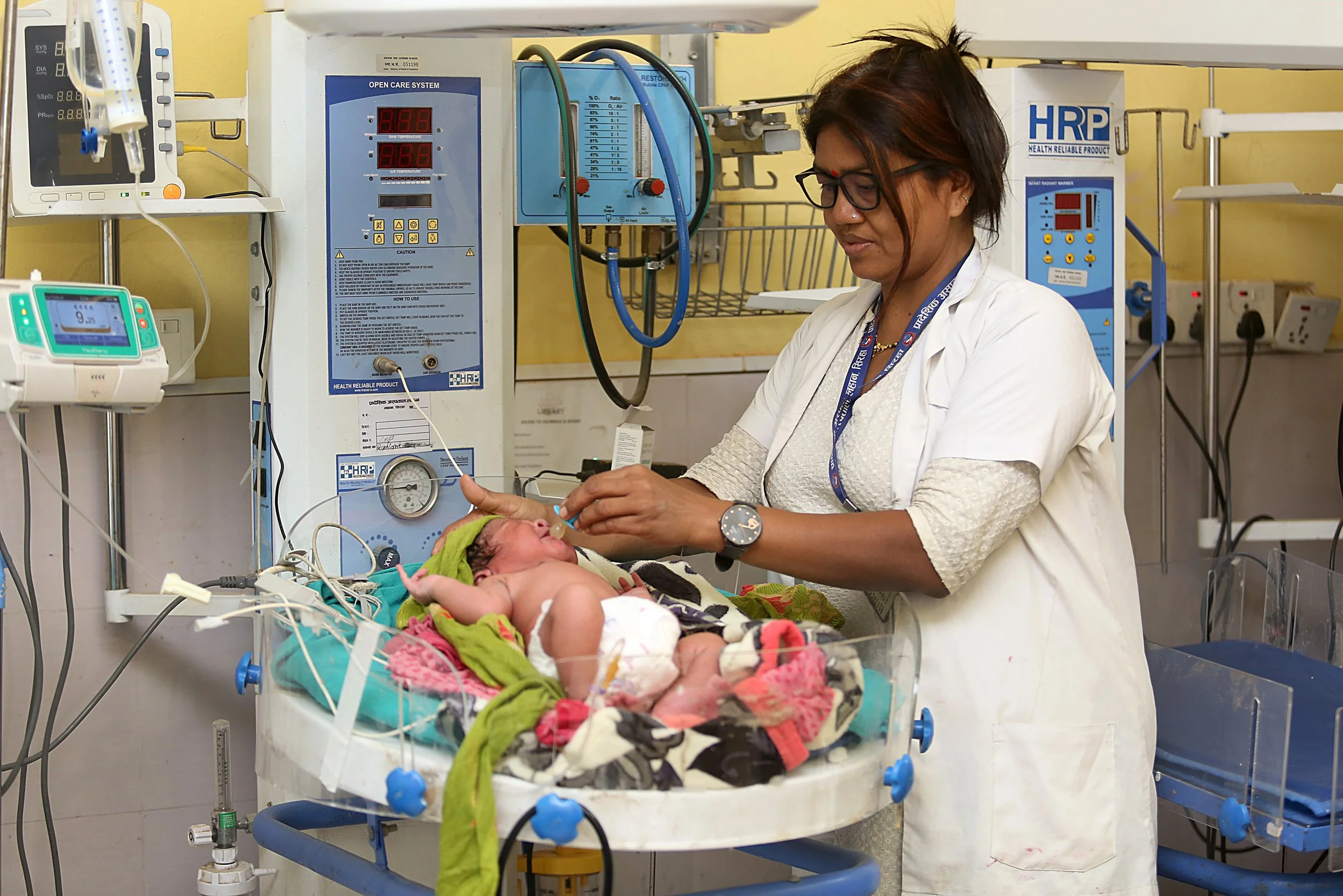 A health worker tends to a newborn in the Special Newborn Care Unit at Lahan Hospital, Nepal. The infant lies on an open care warmer surrounded by monitoring equipment, while the provider performs a clinical assessment.