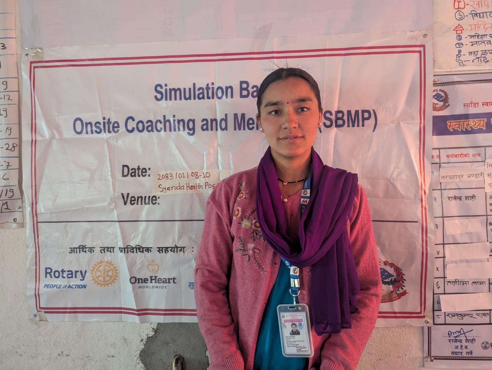 Laxmi Bohara, Auxiliary Nurse Midwife, Syada Health Post in Simkot Rural Municipality, Humla District, stands in front of a banner for a Simulation-Based Onsite Coaching and Mentorship Program (SBMP) session, supported by Rotary and OHW