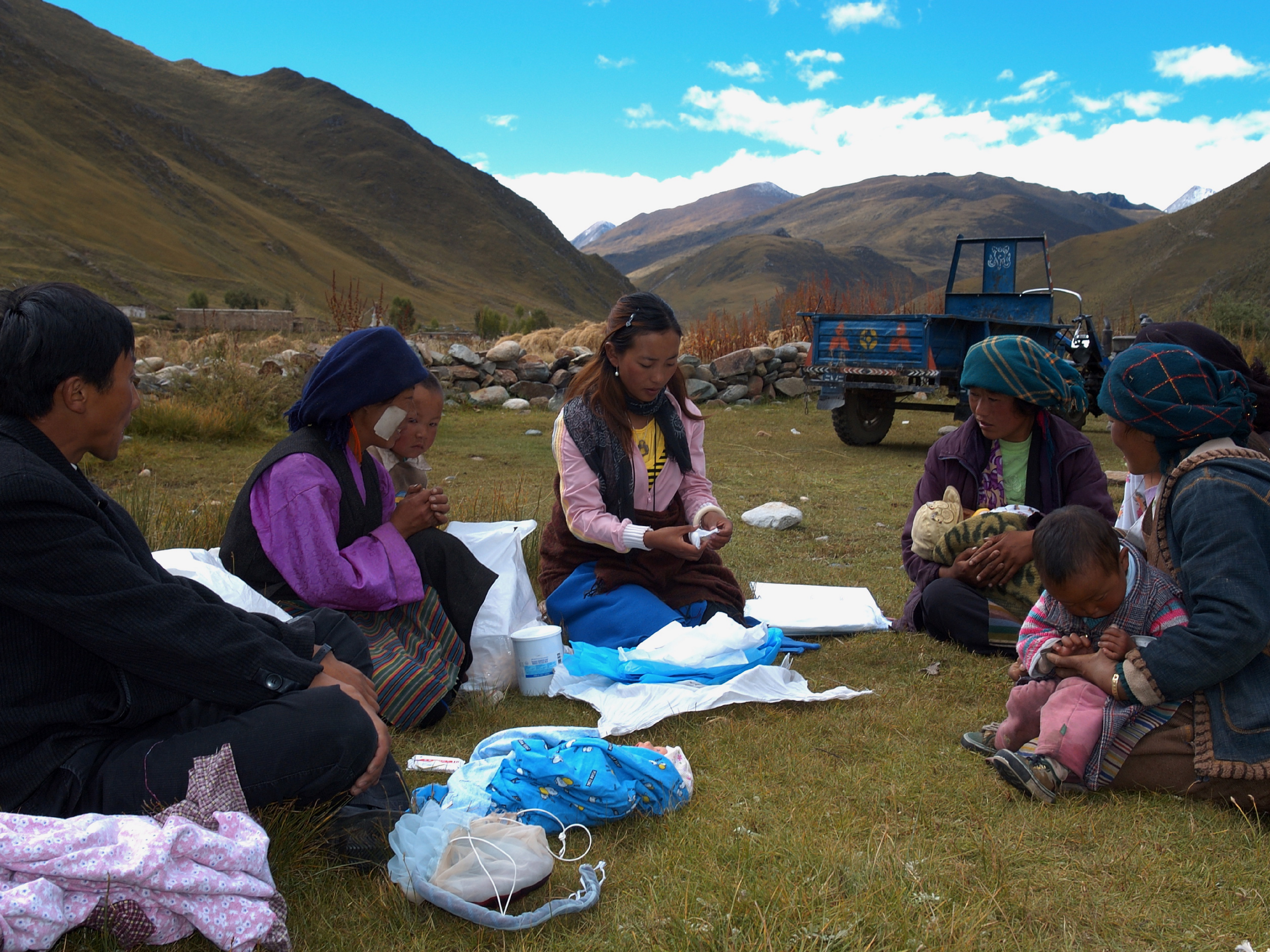A community health session outdoors in a mountain valley in Tibet, where a young woman shows birth supplies to several parents and children seated on the grass.