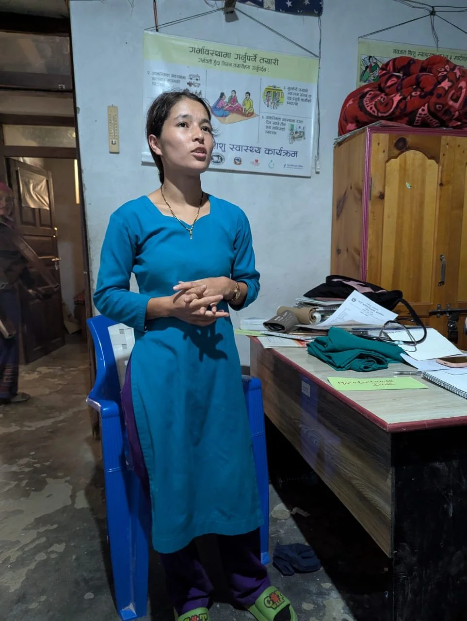 Bindu Mahatara, a staff nurse participating in the Simulation-Based Mentorship Program at Saya Health Post in Humla District, speaks animatedly while seated in her clinic. Birth preparedness and newborn health education posters are displayed