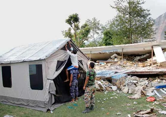 People setting up an emergency, temporary tent that will serve as a birthing center. The background of the photo features rubble of structures damaged in the 2015 earthquakes in Nepal.