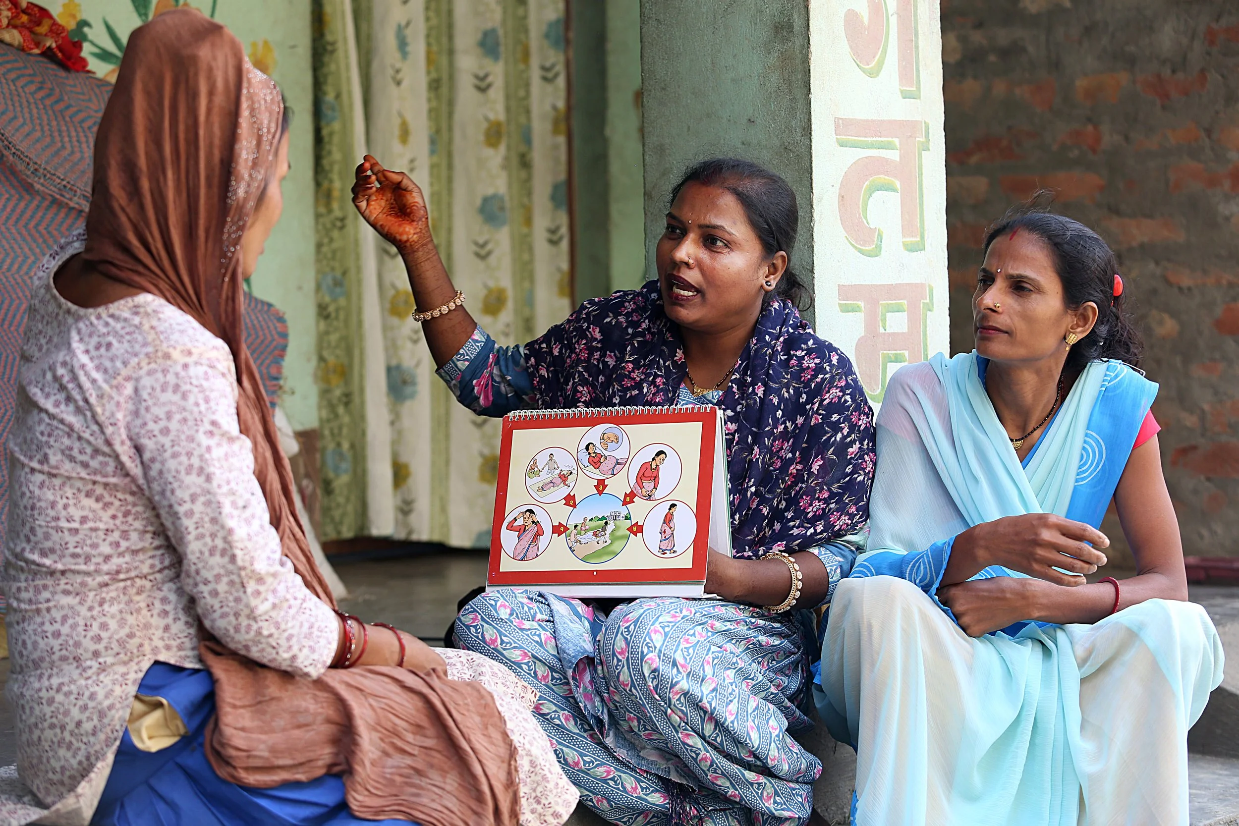 A Female Community Health Volunteer uses an illustrated counseling flip chart to explain maternal health danger signs to two women during a home visit.
