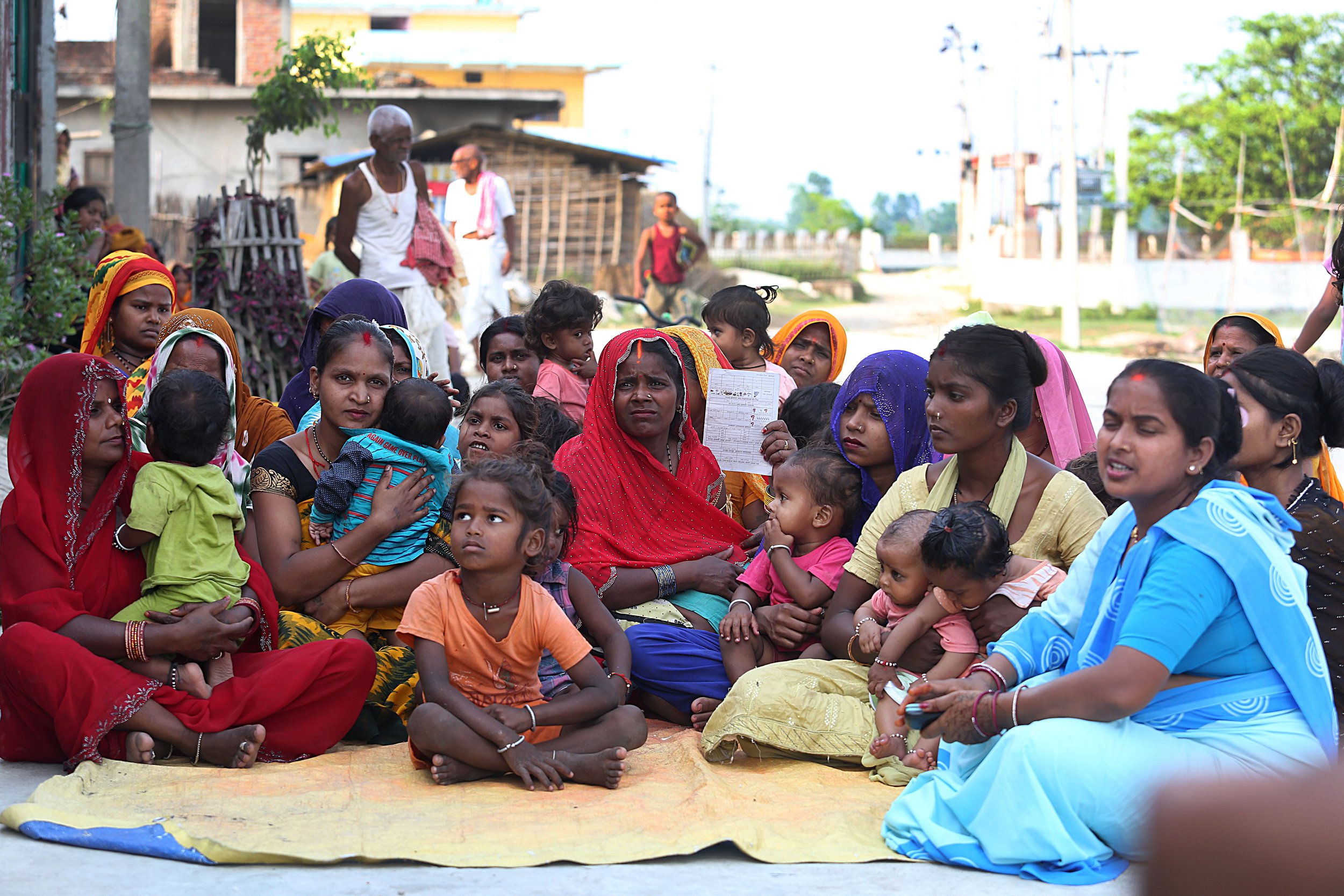 A group of mothers and their children sit together outdoors during a community health session in Nepal, one holding a health record card.