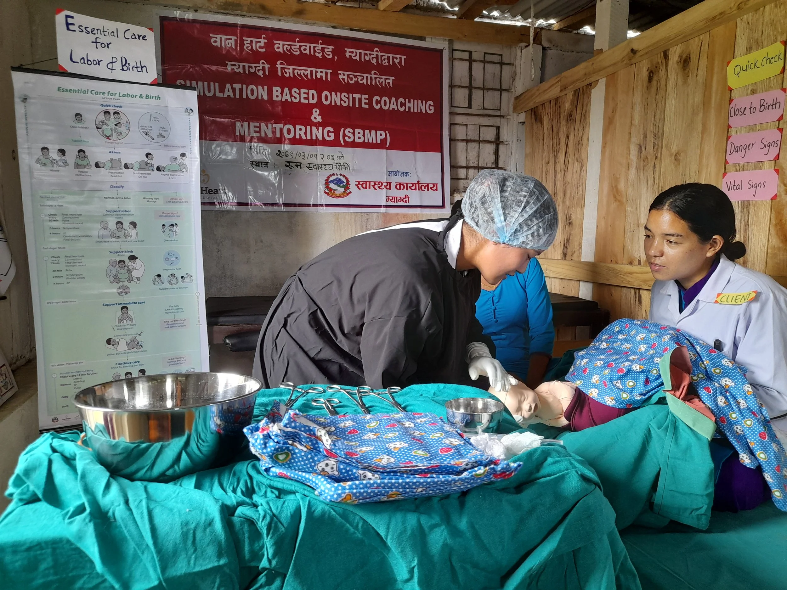 A nurse practices delivery skills on a training manikin during a One Heart Worldwide simulation-based mentoring session in Myagdi District, with a banner and clinical guidelines displayed behind her.