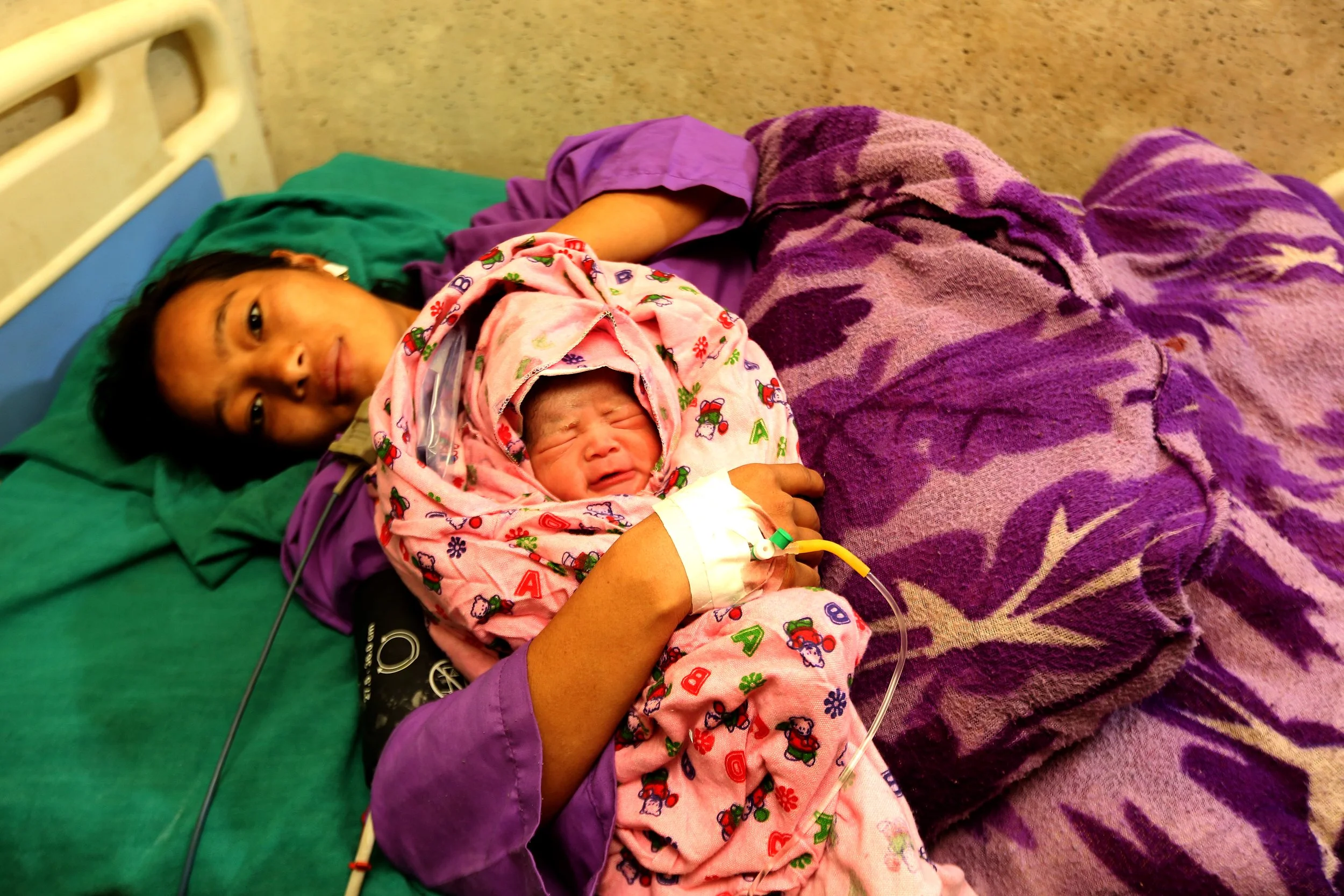 A mother rests in a health facility bed in Nepal, holding her swaddled newborn shortly after delivery.