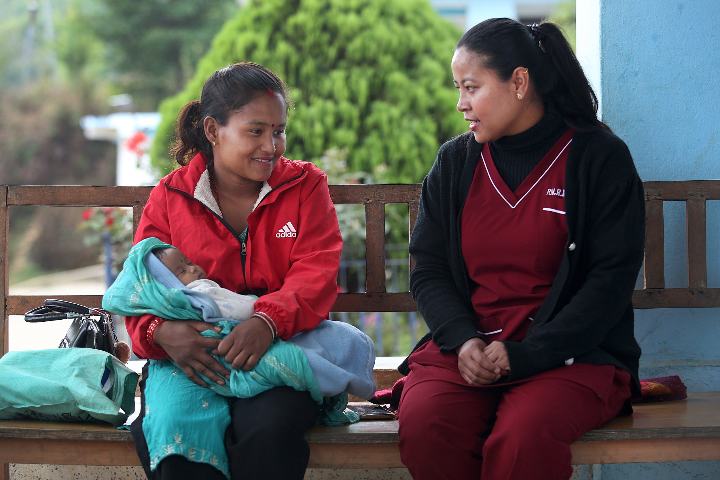 A new mother holds her swaddled baby and talks with a nurse on a bench outside a health facility in Nepal.