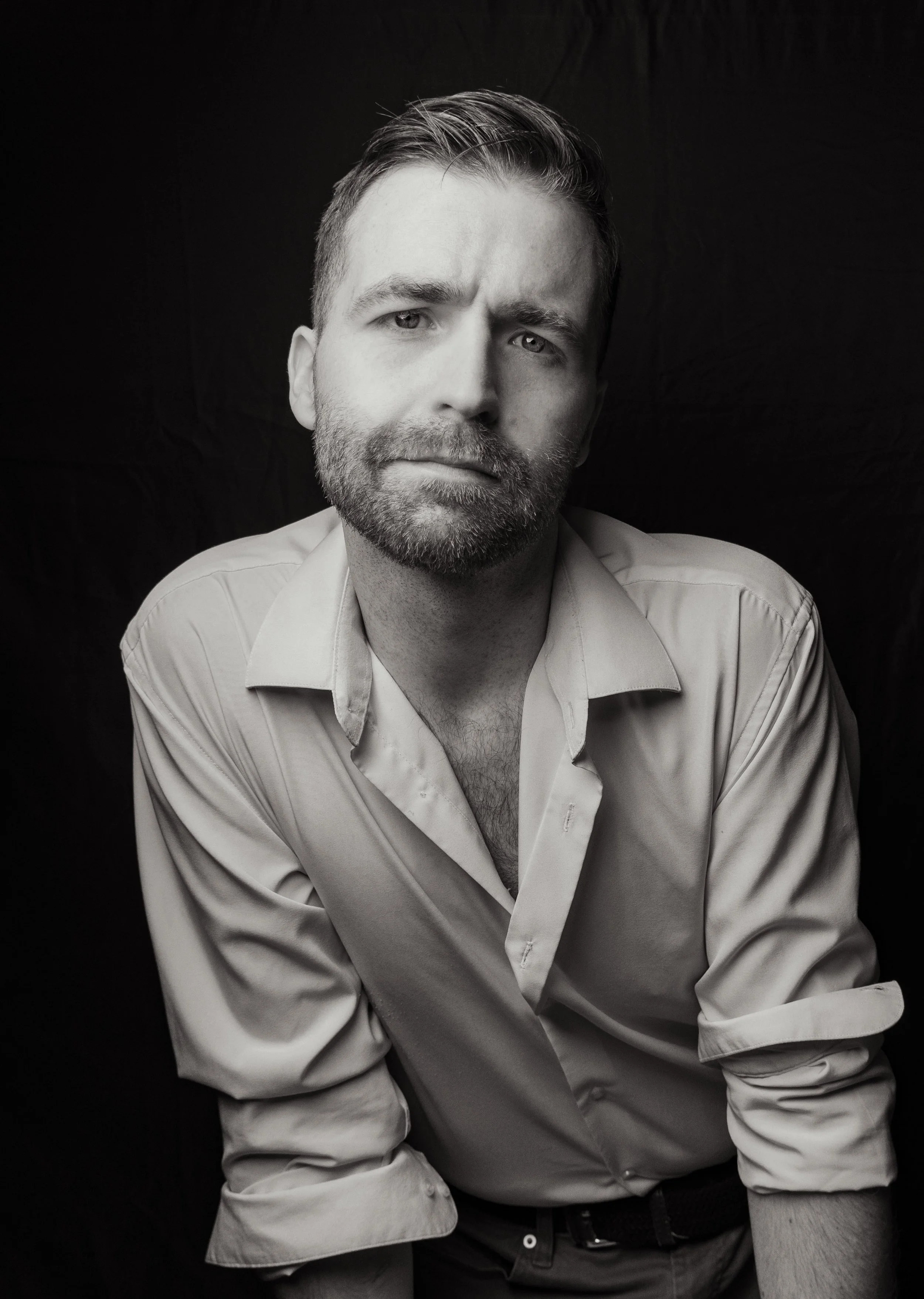 Black and white portrait of a man with short, styled hair, a beard, wearing a light-colored, partially unbuttoned shirt, against a dark background.