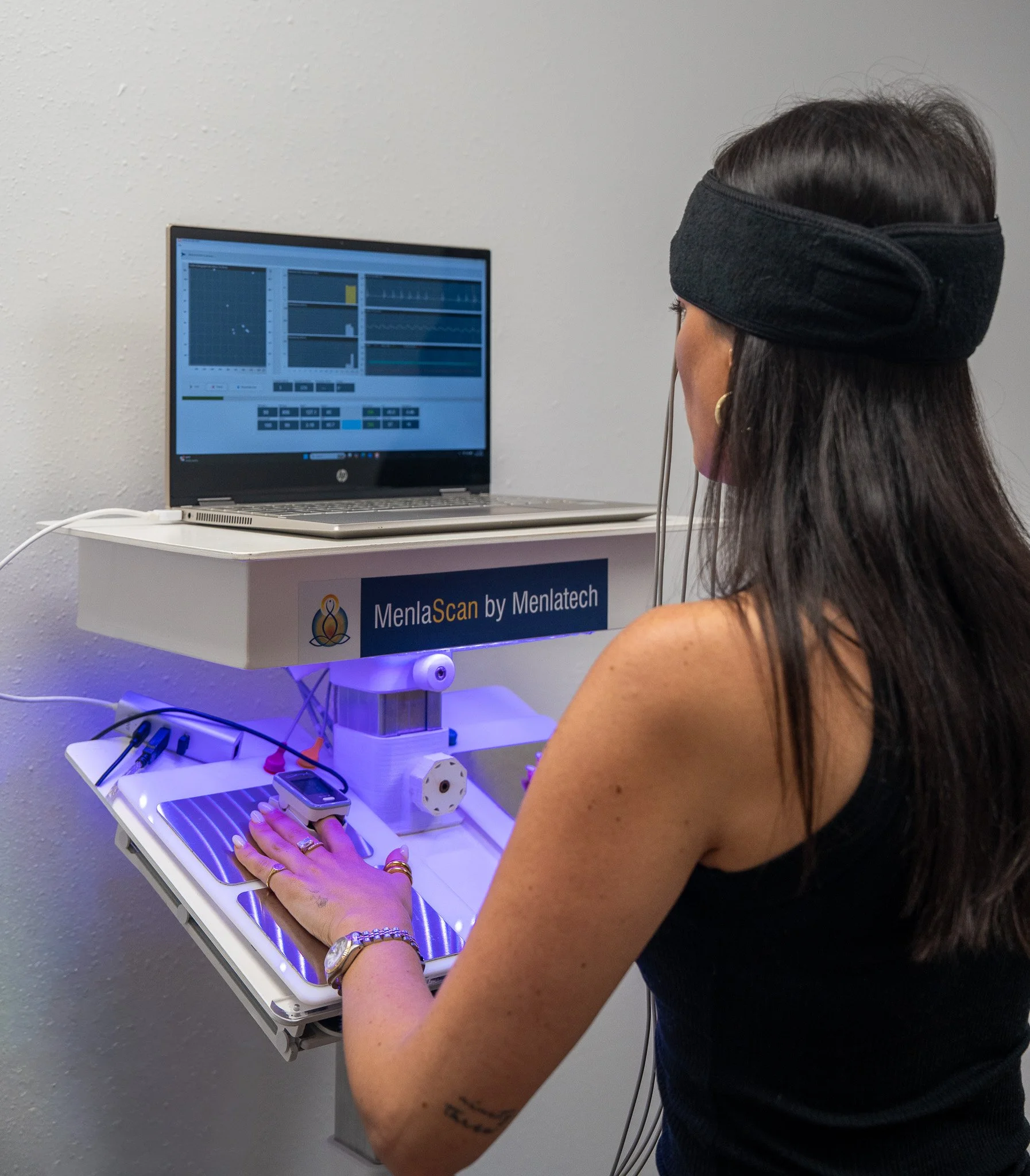 A woman with long dark hair, wearing a black headband and a sleeveless black top, is using her right hand to operate a MenlaScan device with a touchscreen interface. The device is mounted on a white stand labeled 'MenlaScan by Menlatech', and the woman is wearing a gold watch, a gold bracelet, and a gold ring. The room has a plain white wall as background and various connected cables and electronic components are on the shelf below the device.