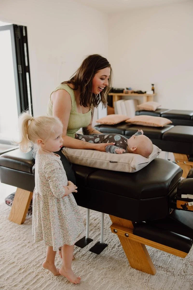 Chiropractor adjusting newborn on support pillow while toddler sister watches from beside the table — pediatric chiropractor in Tulsa, Oklahoma