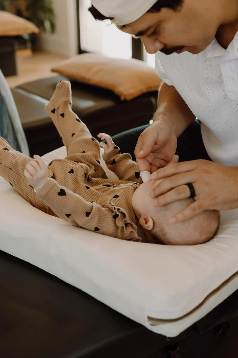 Chiropractor performing gentle fingertip adjustment on newborn in brown-print onesie — infant chiropractic care at Free Spirit Chiropractic in Tulsa