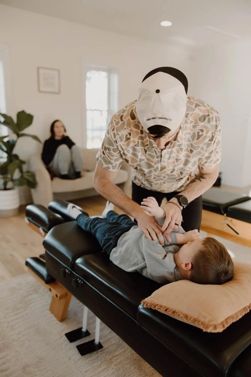 Dr. Colten in cap performing gentle adjustment on young child on table with mom seated nearby — pediatric chiropractor in Tulsa, Oklahoma