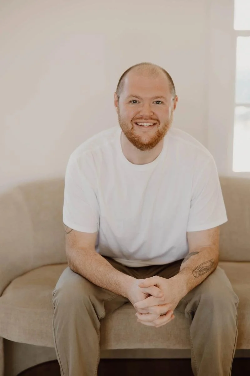 Dr. Jake Jones portrait seated on beige sofa wearing white t-shirt, red beard visible — Free Spirit Chiropractic of Tulsa, Oklahoma