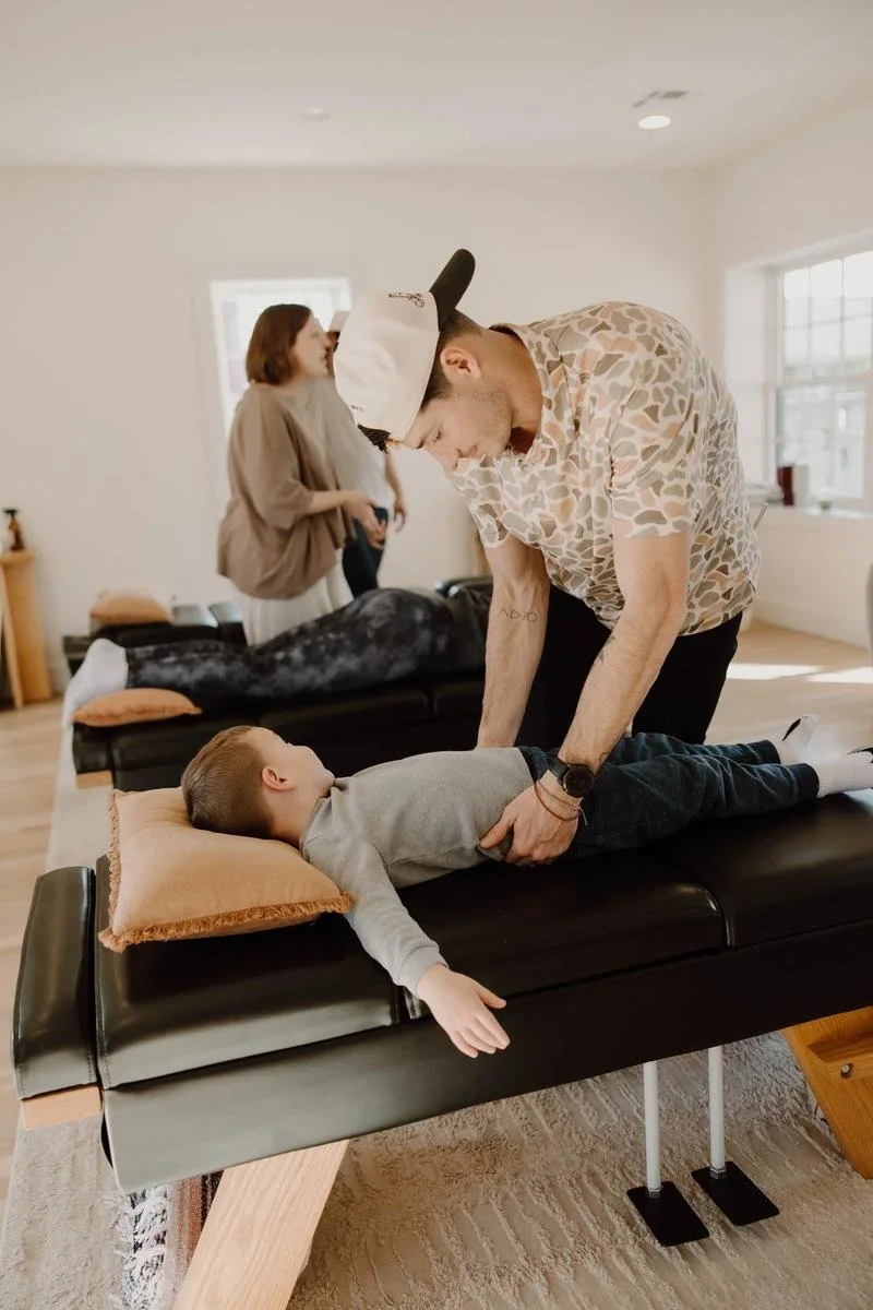 Dr. Colten in cap performing gentle adjustment on young child while parent holds infant in background — pediatric chiropractor in Tulsa, Oklahoma